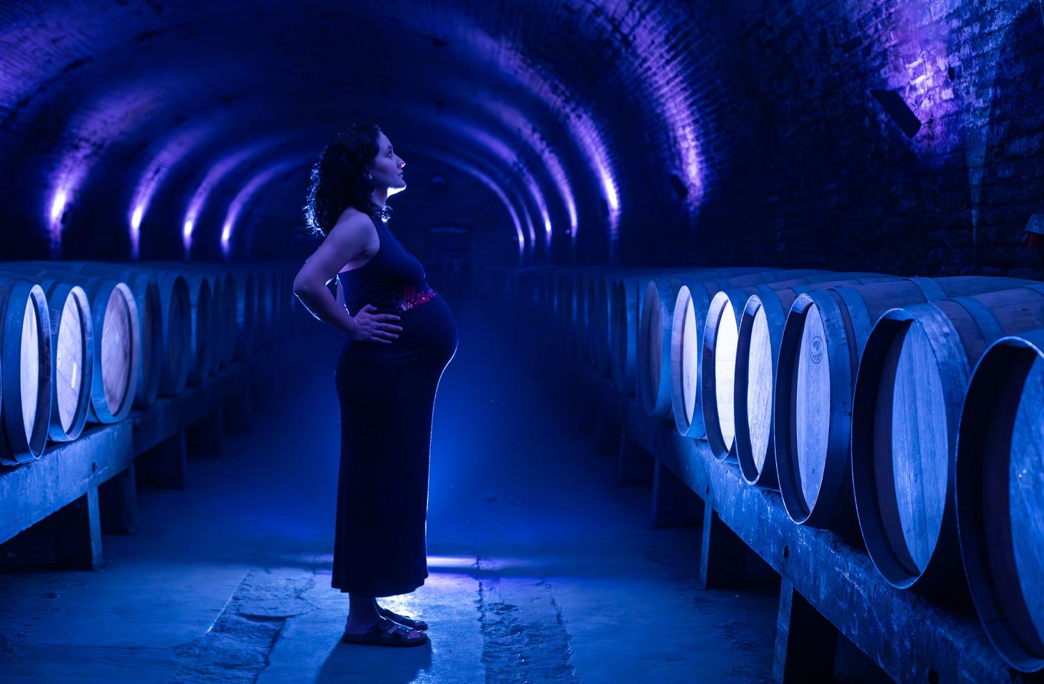 Pregnant woman in black dress poses in blue-lit arched wine cellar with wooden barrels.