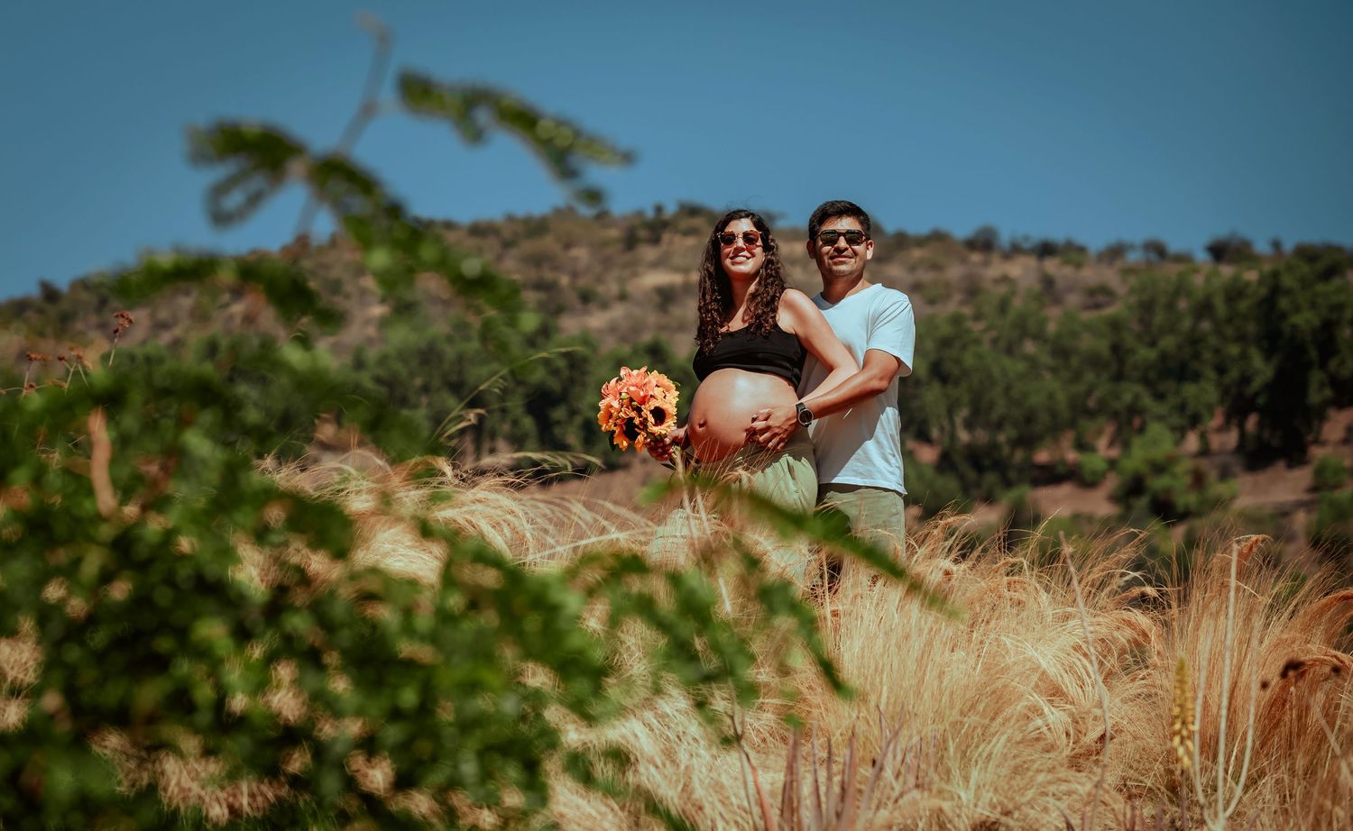 Pregnant couple embracing in a golden field with hills, woman holding a colorful bouquet under blue sky.
