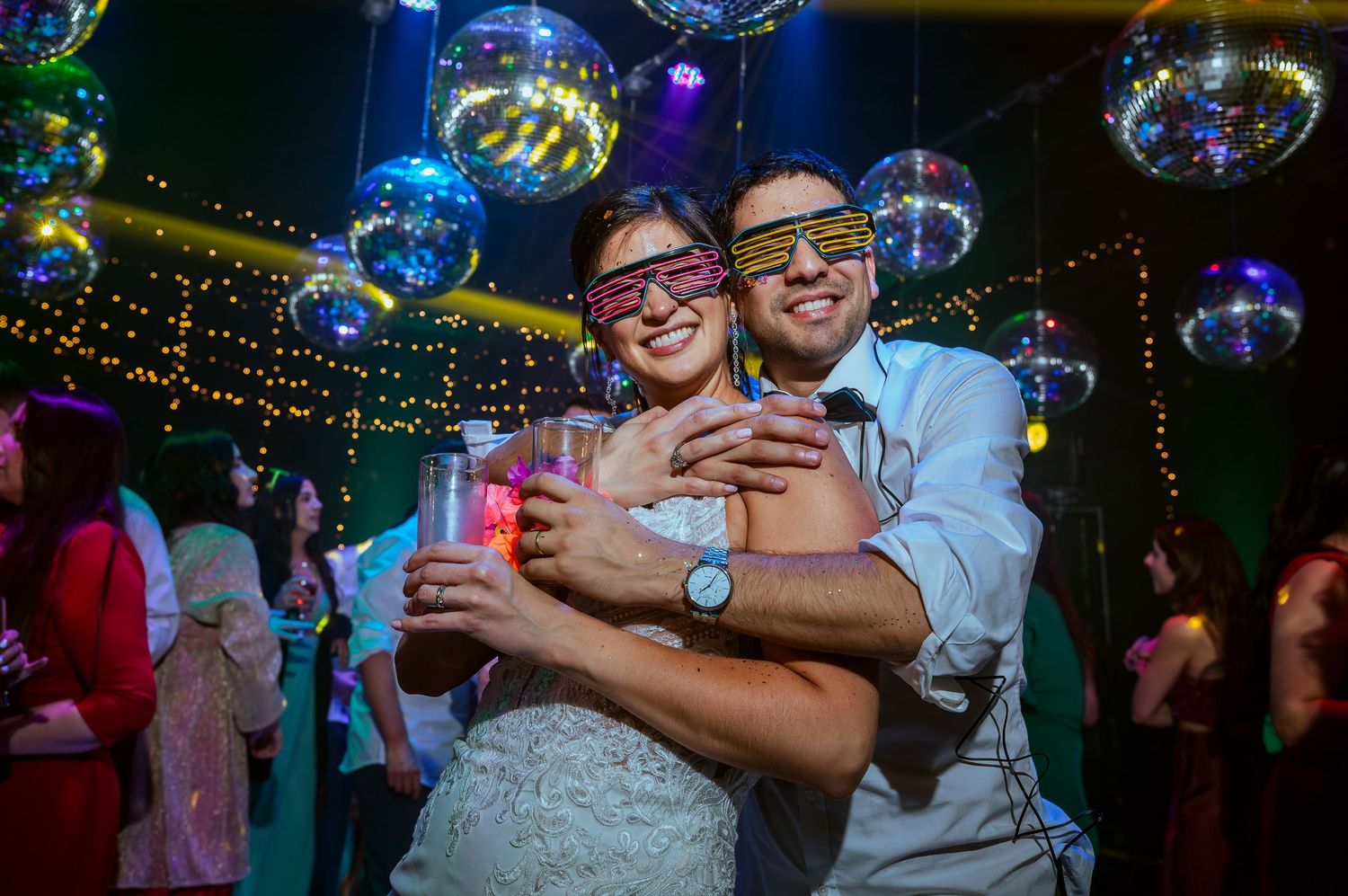 Bride and groom wearing colorful neon glasses dancing together at a festive wedding reception with disco ball decorations.