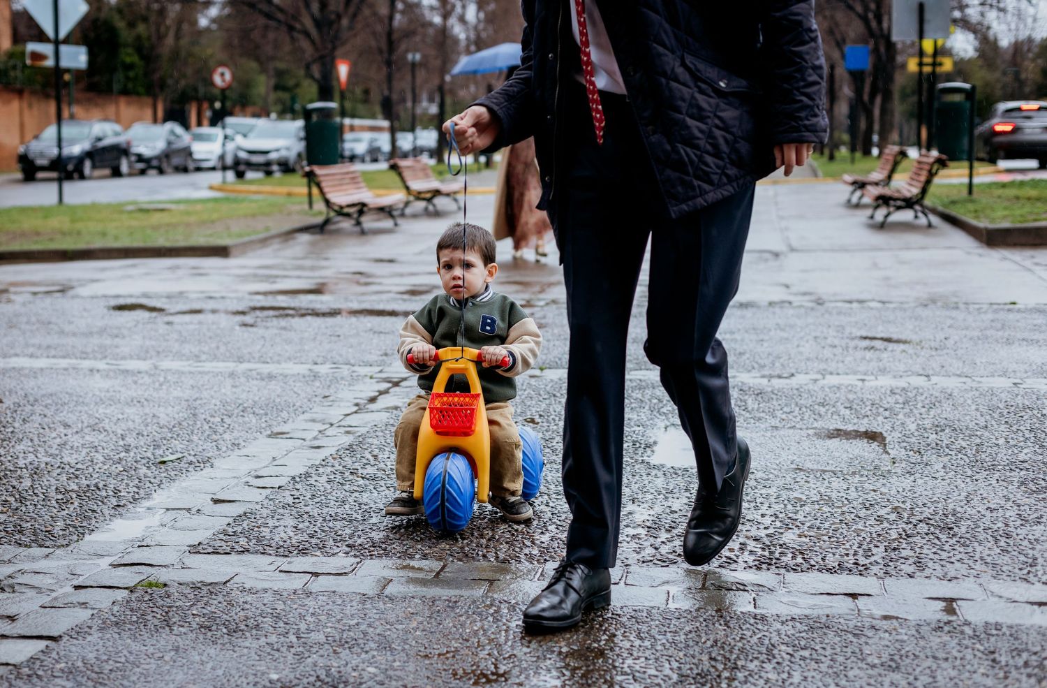 Adult guiding a young child on a colorful toy tricycle on a wet rainy day sidewalk in a park.