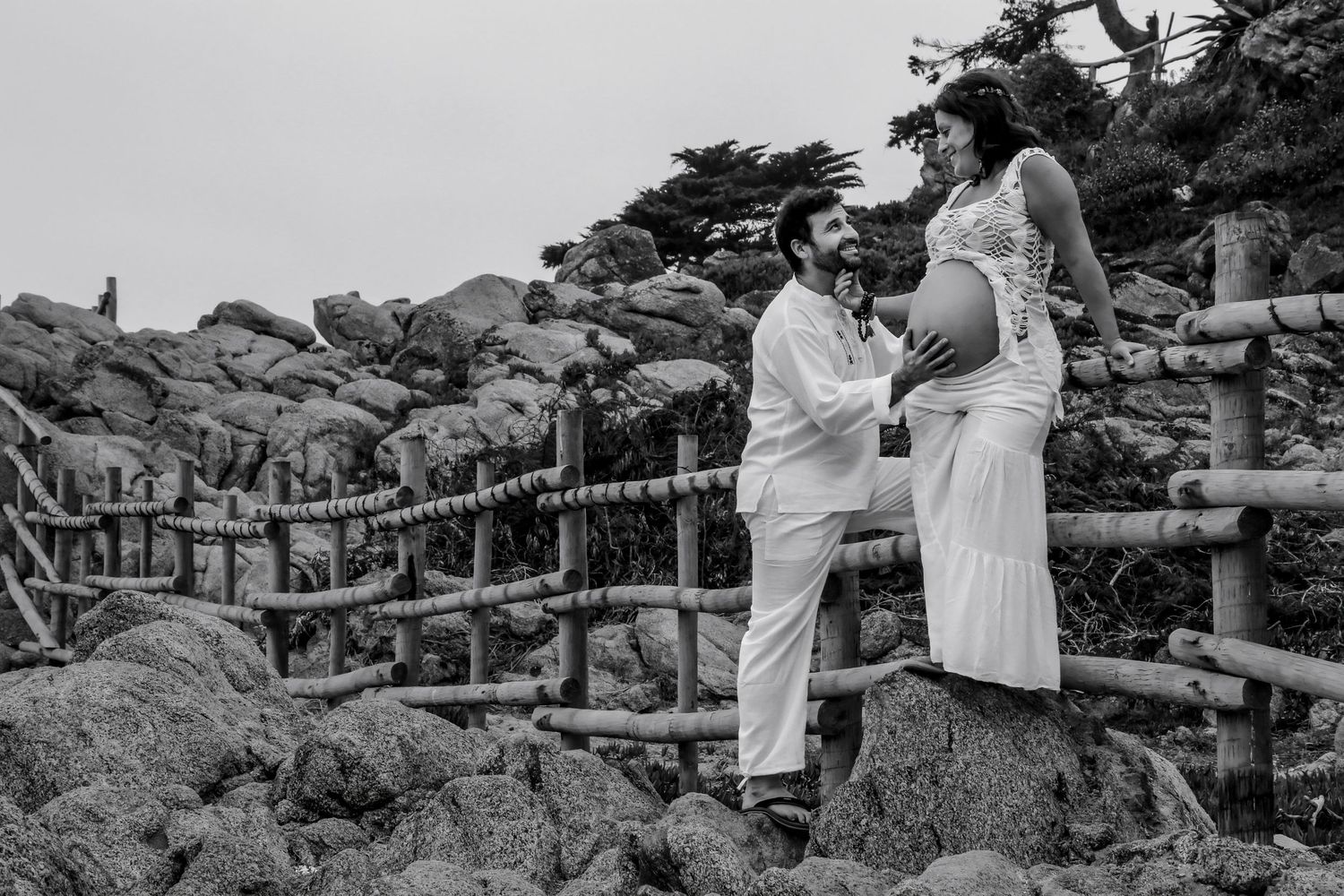 Black and white maternity photo of a couple on a rocky beach, man touching pregnant woman's belly by a bamboo fence.