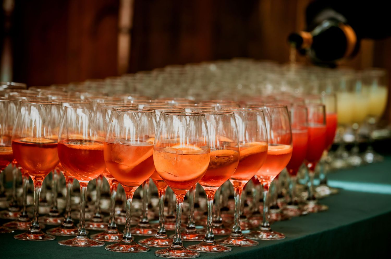 Rows of elegant wine glasses filled with pink Aperol Spritz cocktails arranged on a green table at a catered event.