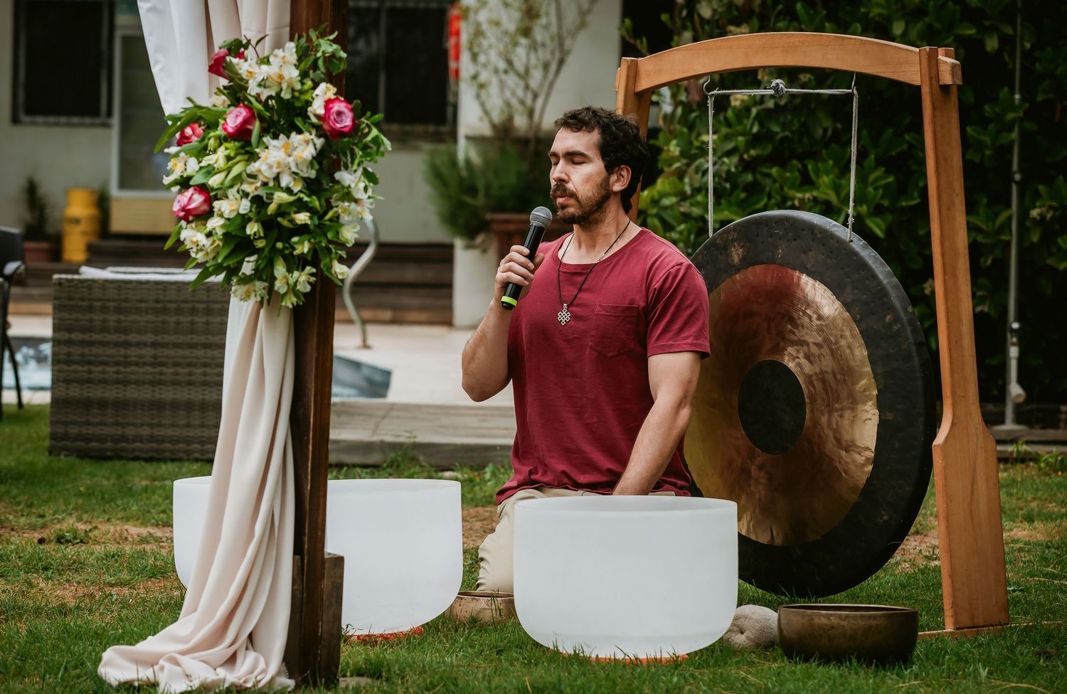 Man in red shirt speaks into microphone near large gong and white crystal bowls at outdoor wellness event.