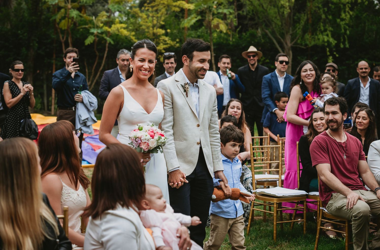 Bride and groom walk down the aisle at an outdoor wedding ceremony surrounded by smiling guests in a garden setting.