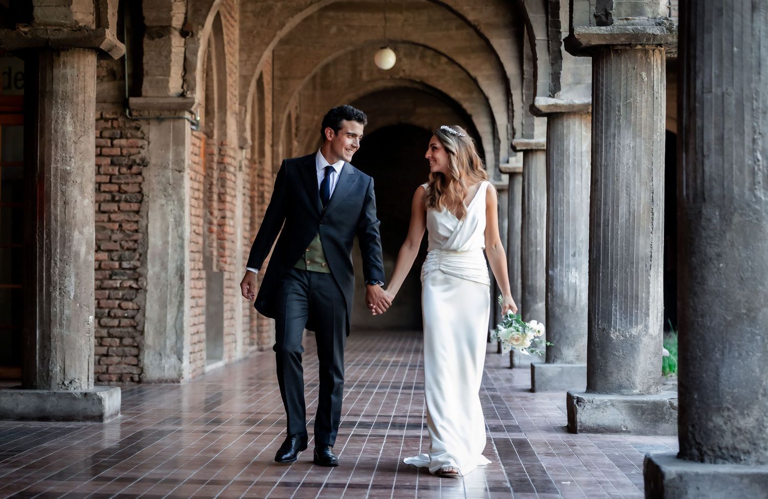 Bride and groom walking hand in hand under elegant arched colonnade at historic wedding venue.