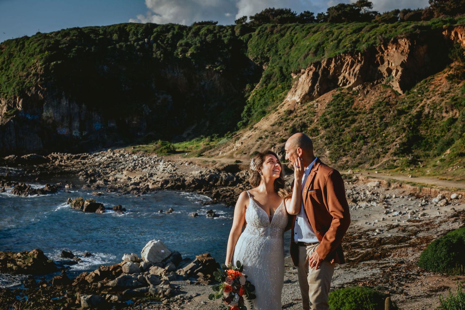 Bride in a white lace gown holding a bouquet embraces her groom in a rust jacket on a rocky coastal beach with green cliffs behind.