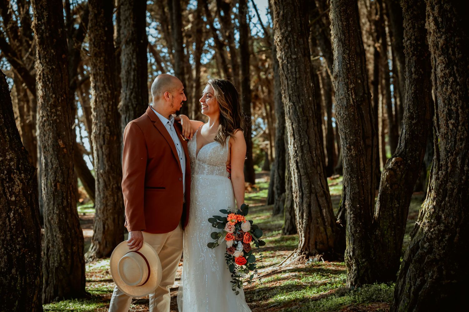 Bride in a white lace gown holding a colorful bouquet embraces a groom in a rust blazer holding a straw hat among tall forest trees.
