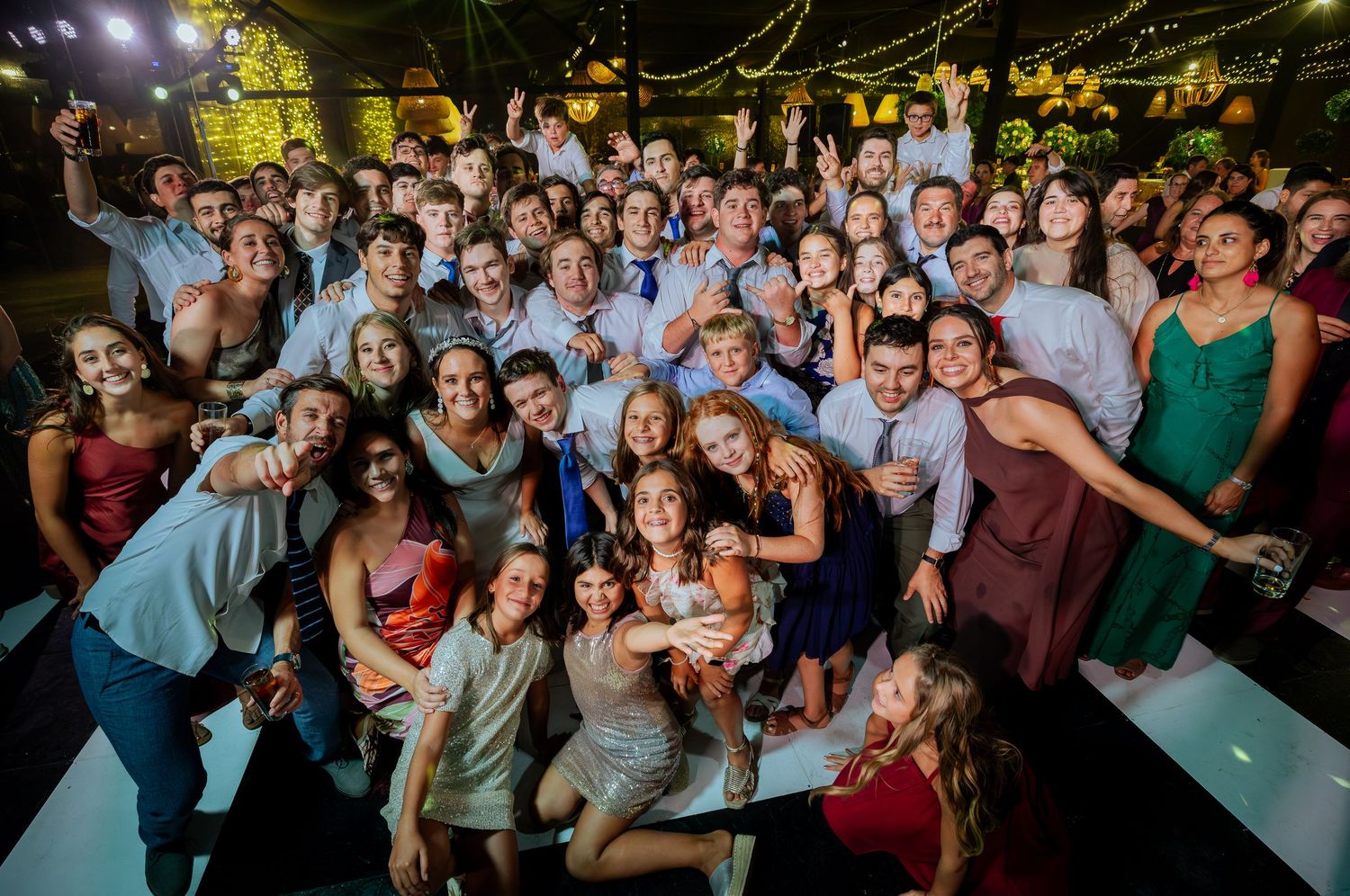 Large crowd of wedding guests smile and pose together on outdoor dance floor at night.