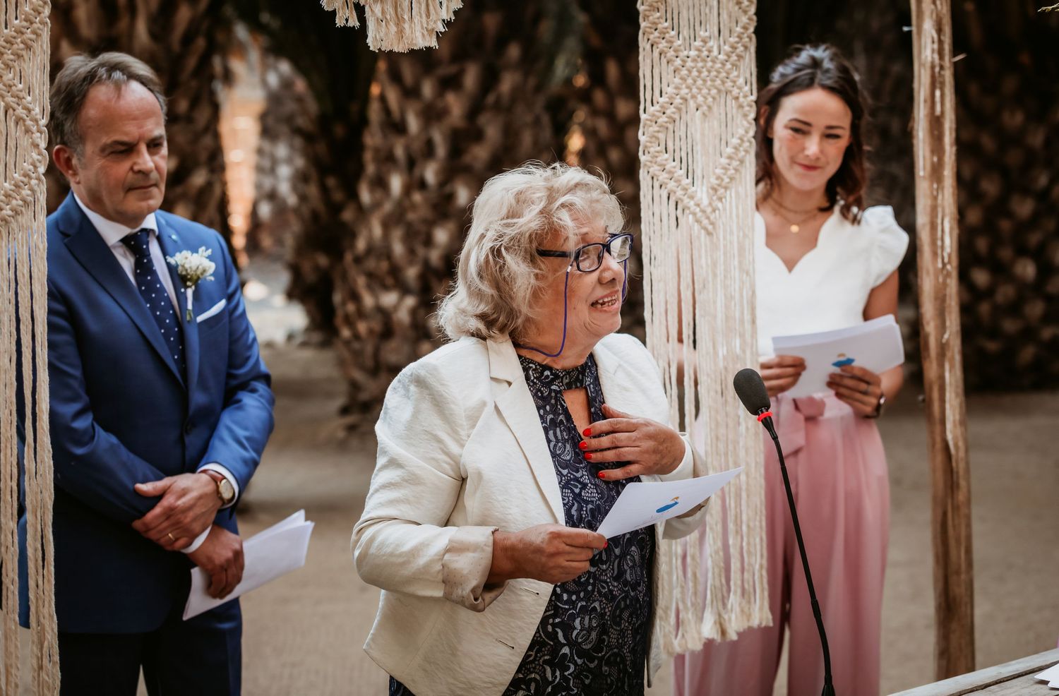 Elderly woman in white jacket delivers speech at boho outdoor wedding ceremony with macrame backdrop.