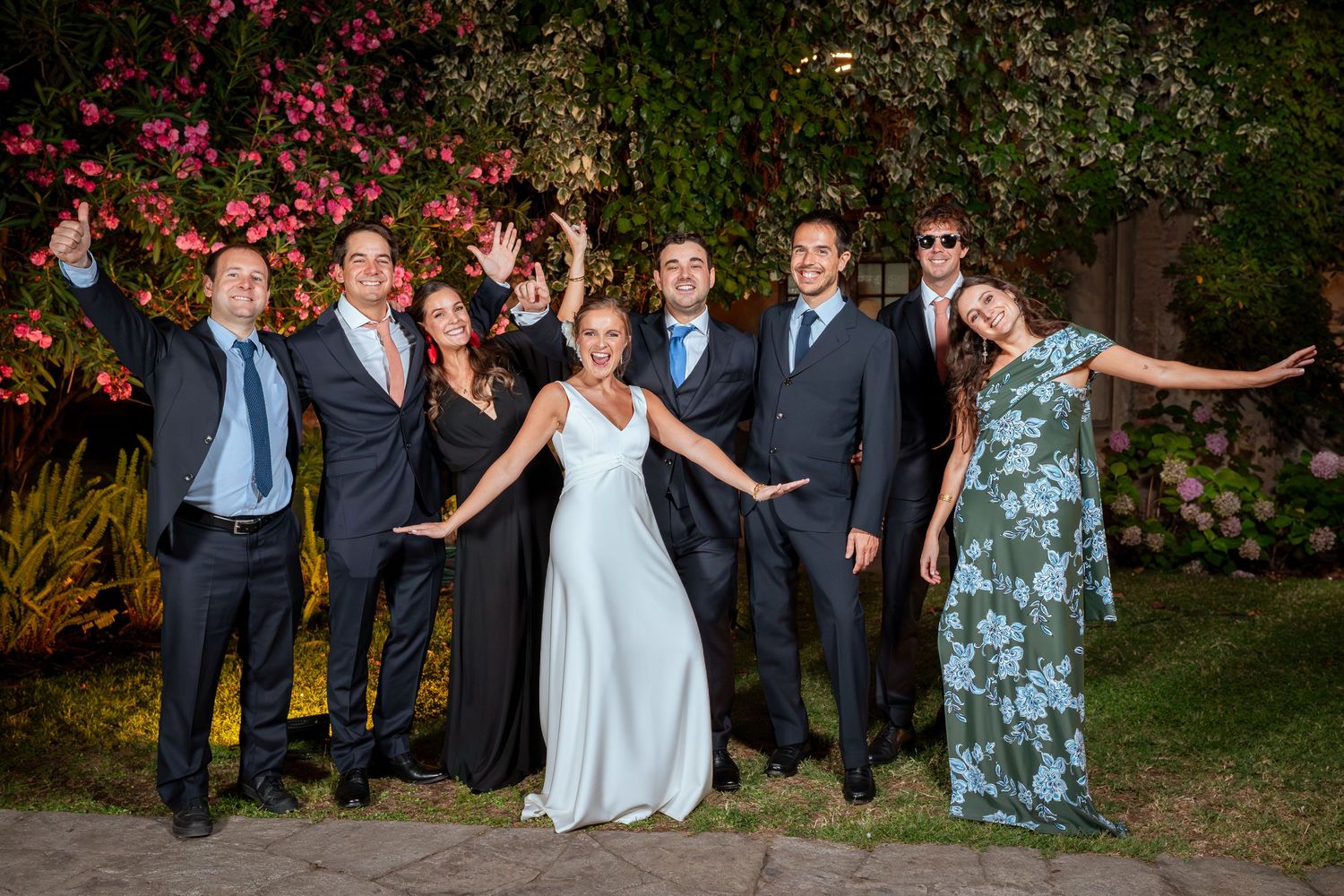 Joyful wedding party group photo with bride and groom among friends in garden setting.