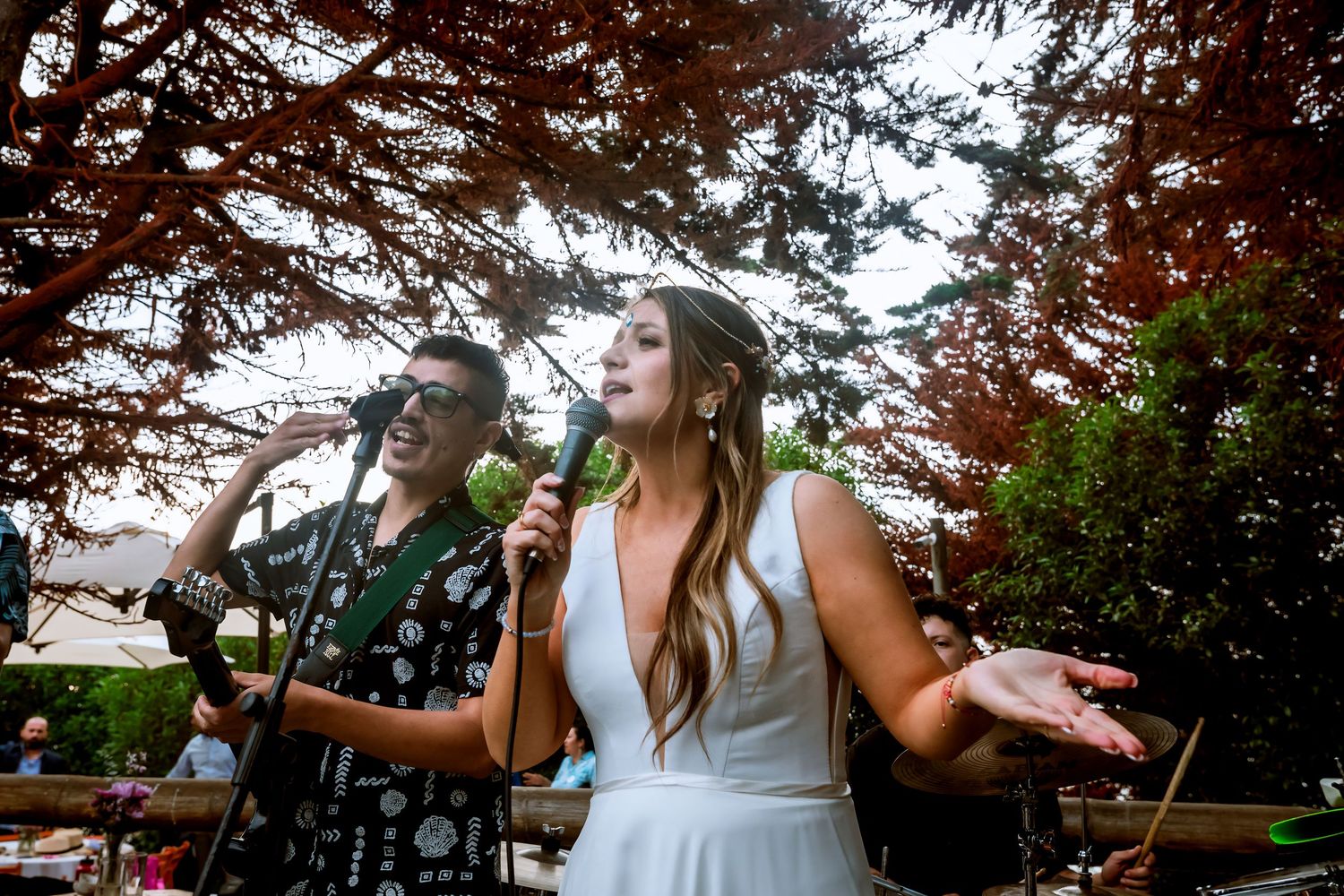 Bride in white dress singing into microphone with band member at outdoor wedding reception.