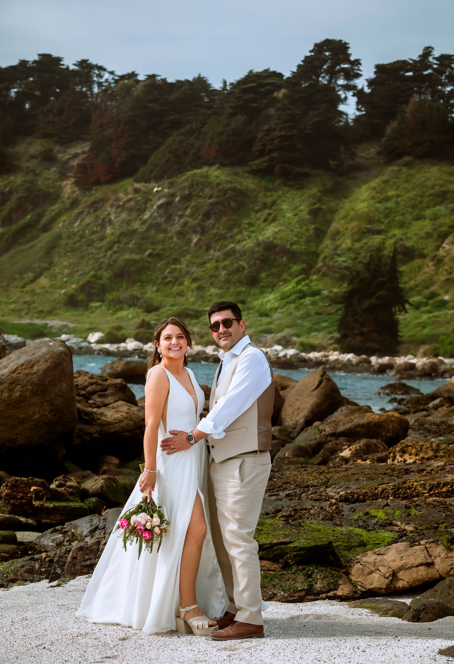Smiling bride and groom posing on rocky coastal beach with lush green hillside and trees in background.