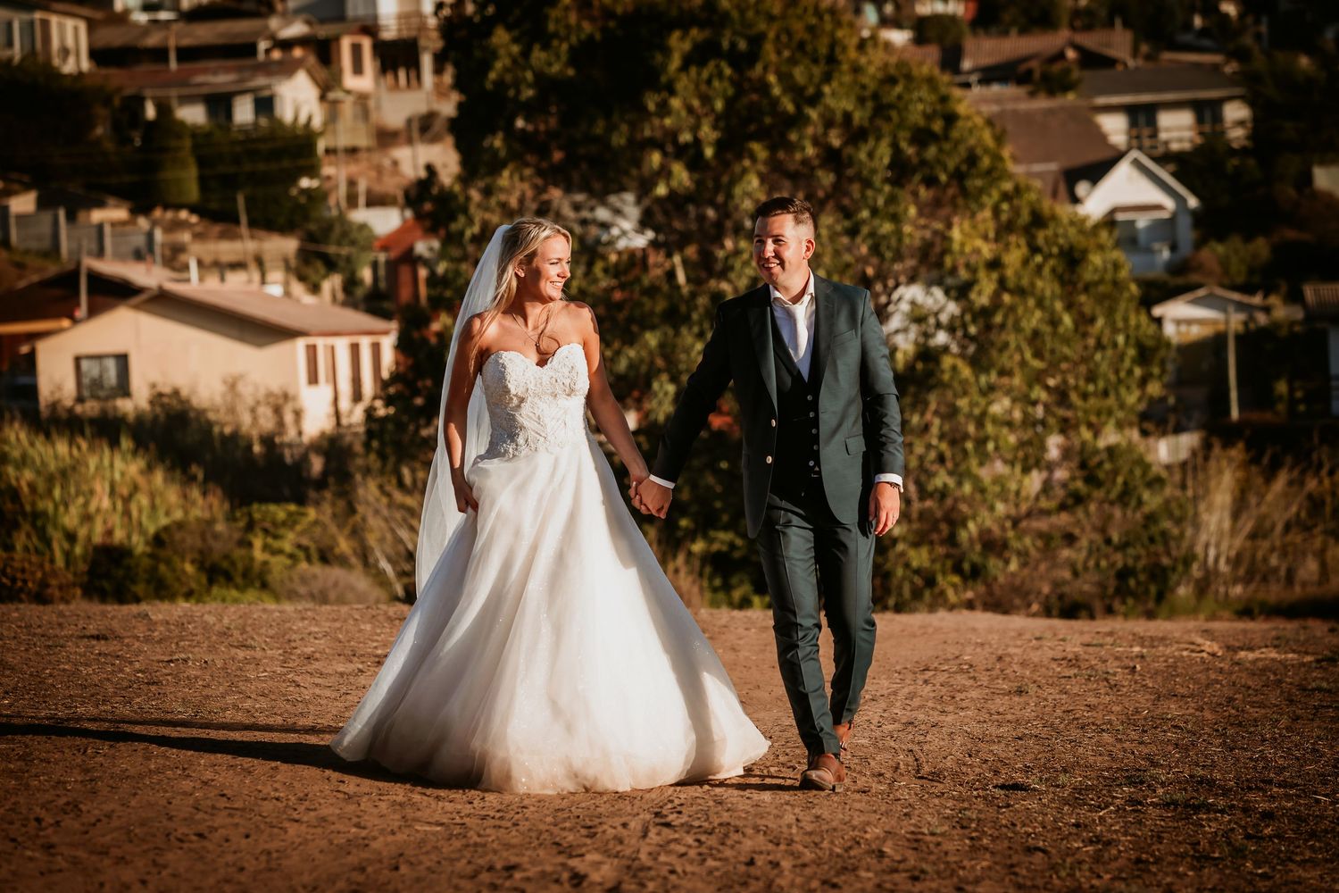 Smiling bride and groom walking hand in hand on a dirt path with town backdrop.