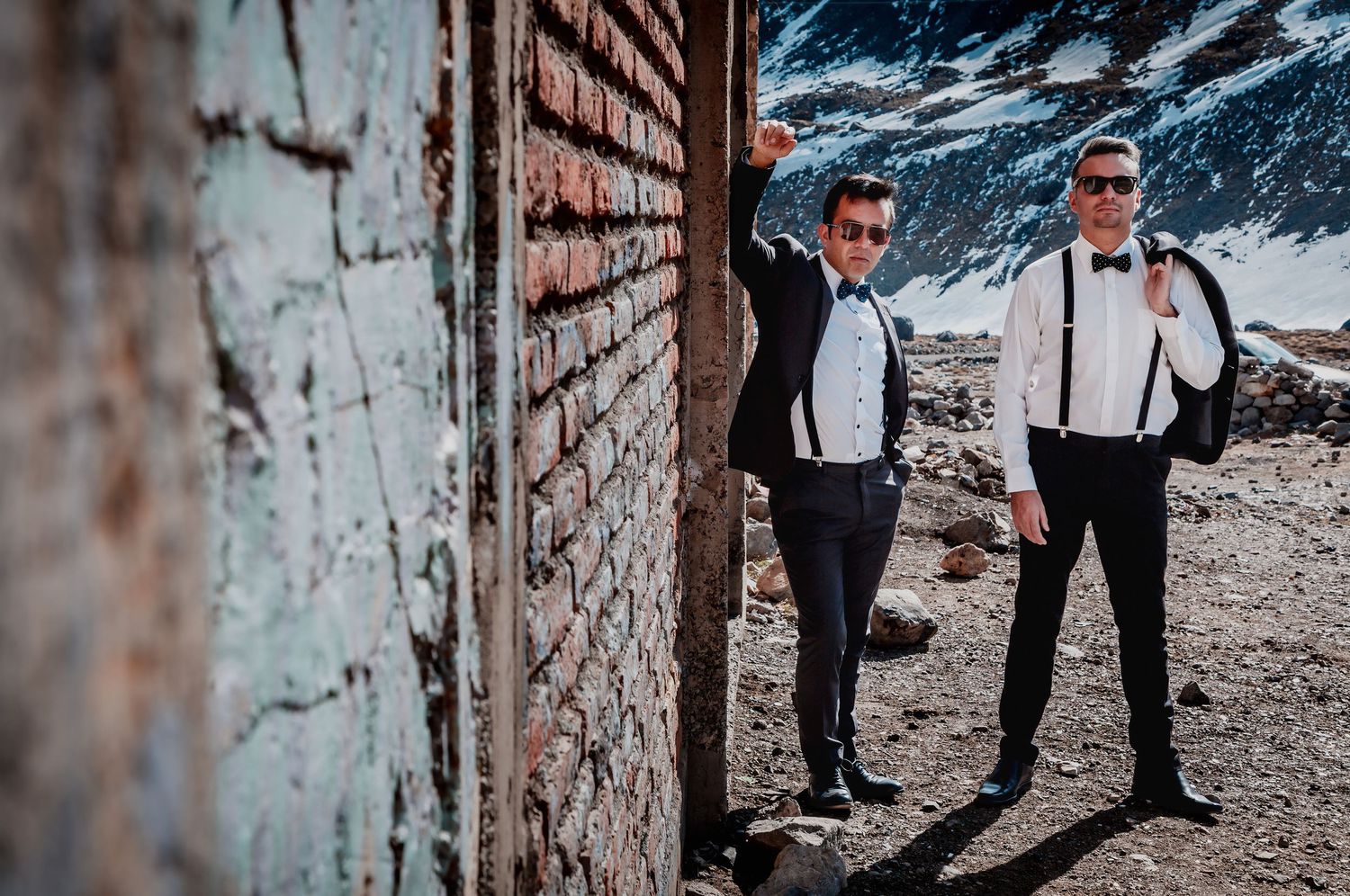 Two grooms in tuxedos with suspenders and bow ties pose by a rustic brick wall in a snowy mountain setting.