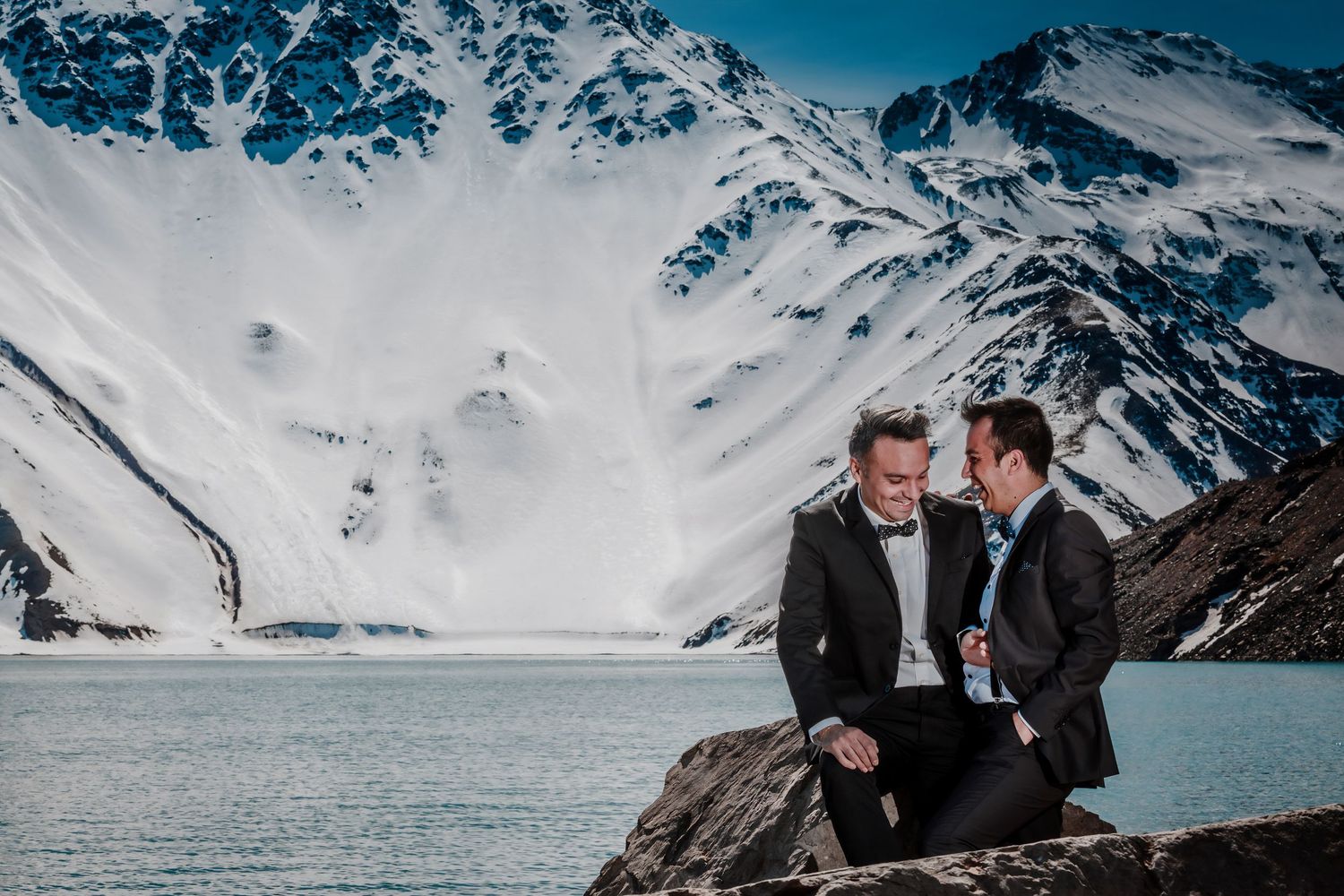 Same-sex couple sits together on rocks by a glacial lake, sharing a quiet wedding moment.
