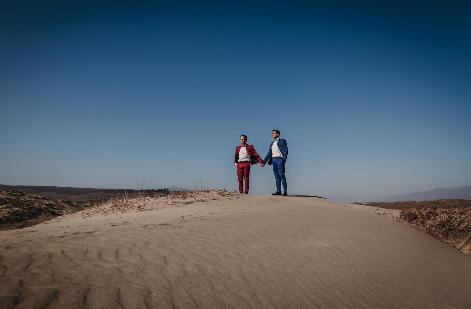 Two men holding hands standing atop a sand dune under a clear blue desert sky.