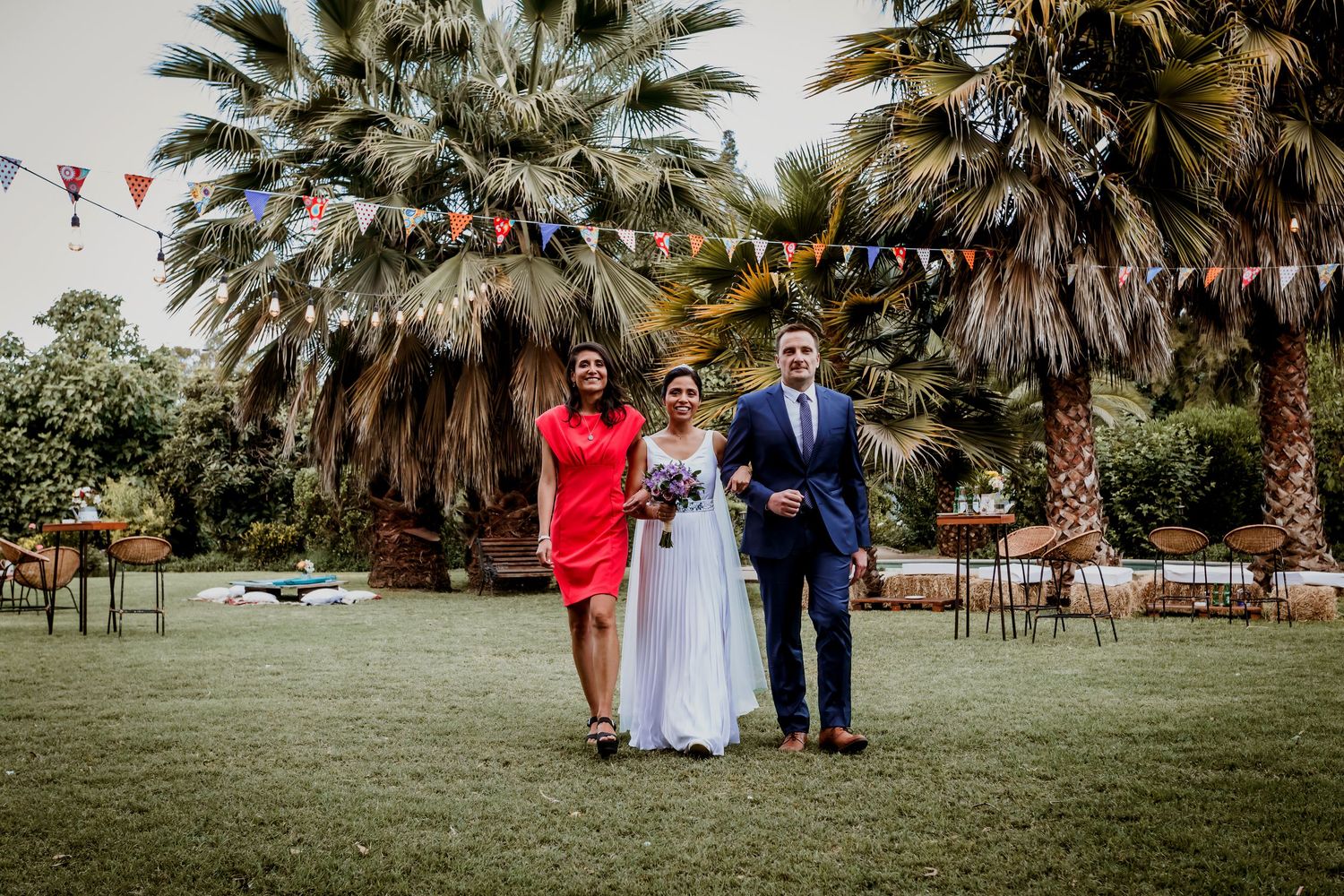 Bride poses with woman in red dress and man in blue suit on lawn with palm trees and bunting.