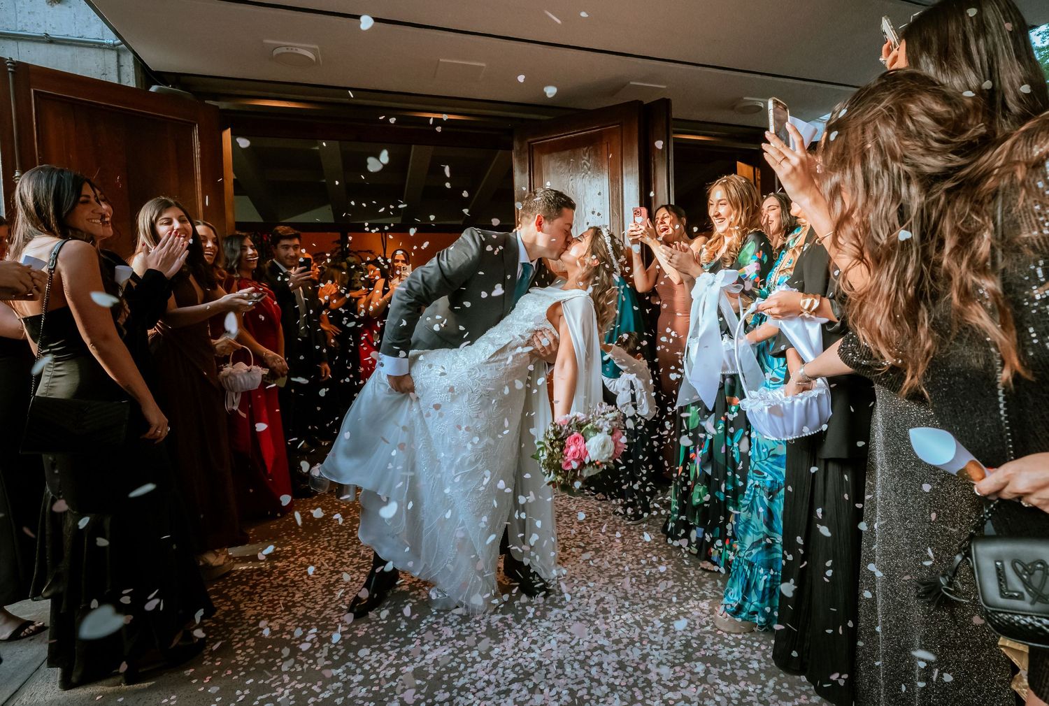 Groom dips bride for a kiss as guests throw white confetti during joyful wedding exit celebration.