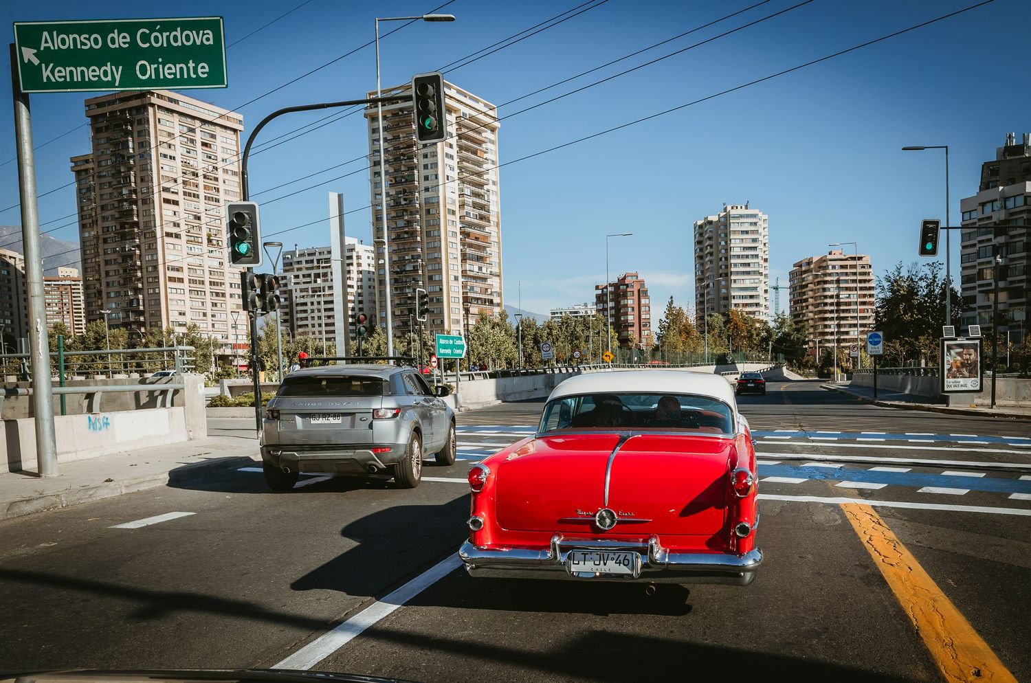 Classic red vintage car driving through a busy city intersection on a sunny day.