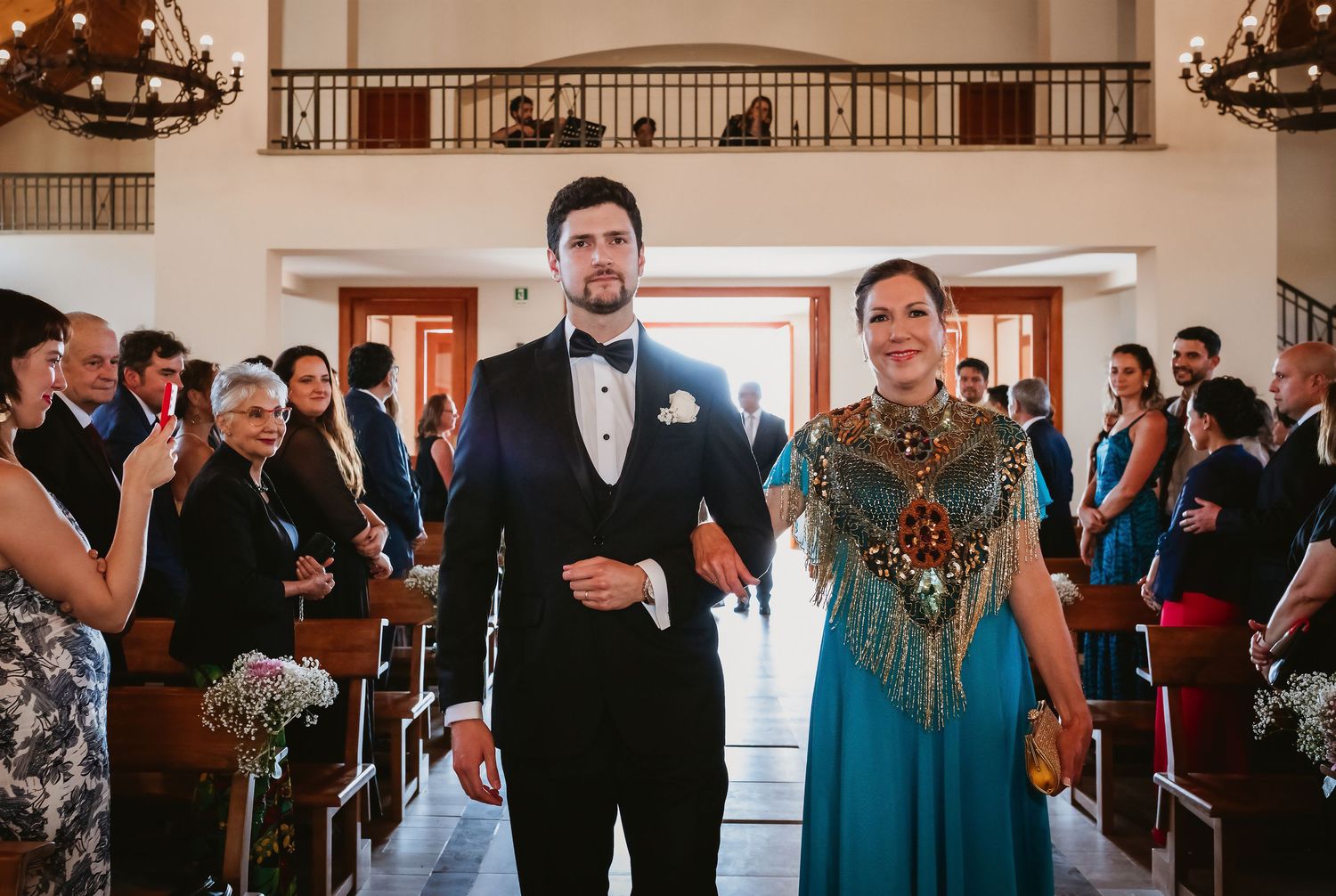 Groom in black tuxedo walking down church aisle with elegantly dressed woman in teal gown.