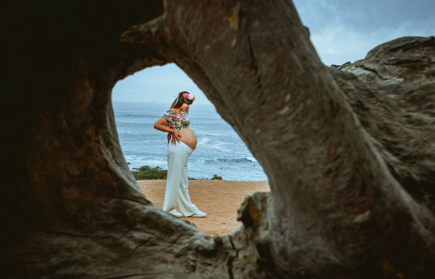 Pregnant woman in floral top and white skirt framed by driftwood arch on beach.