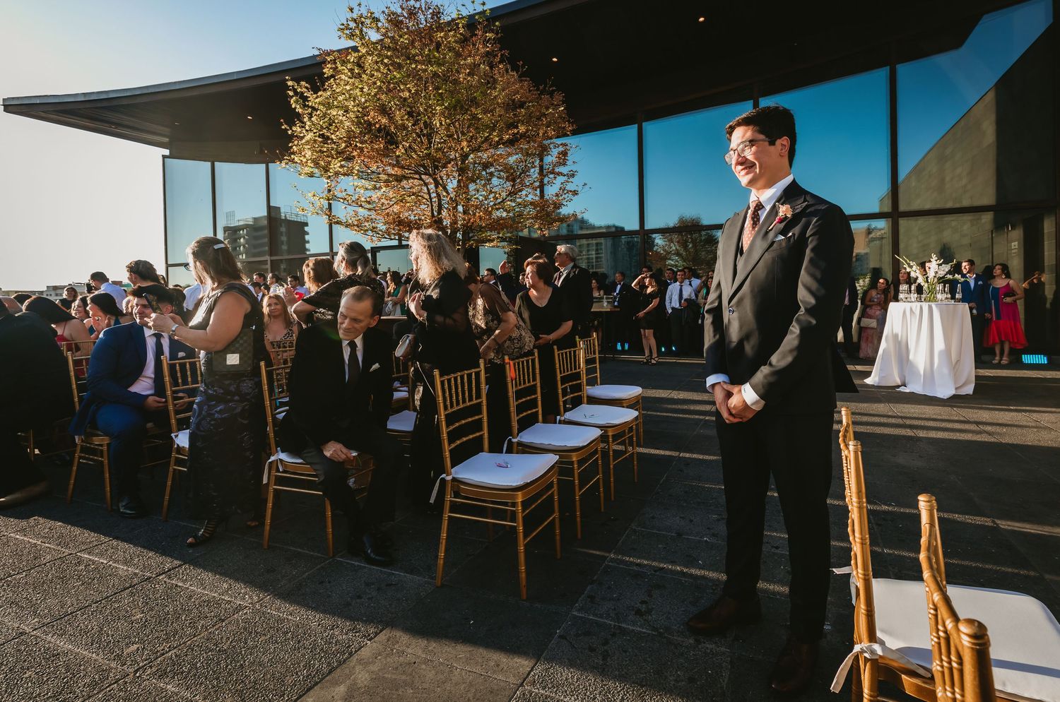 Groom in dark suit stands at end of outdoor wedding ceremony aisle awaiting bride.