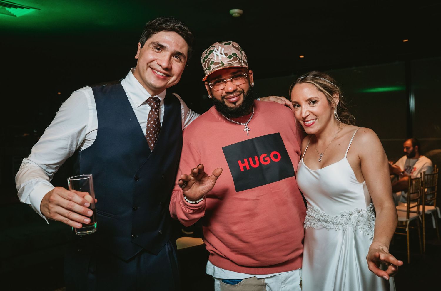 Bride and groom pose with a guest wearing a Hugo Boss sweatshirt at a wedding reception.