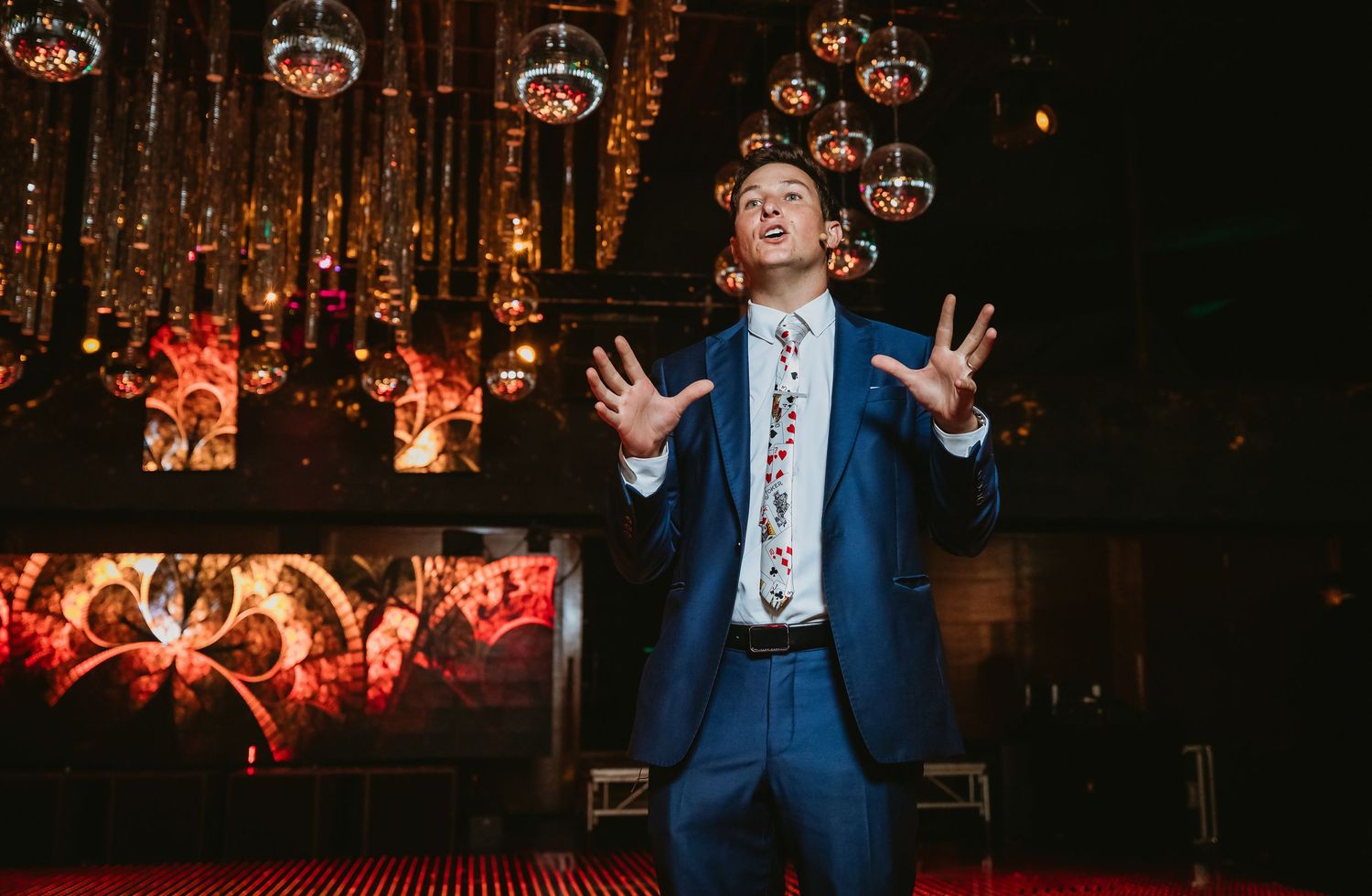 Magician in blue suit performing on stage with dramatic red and gold backdrop at a wedding event.