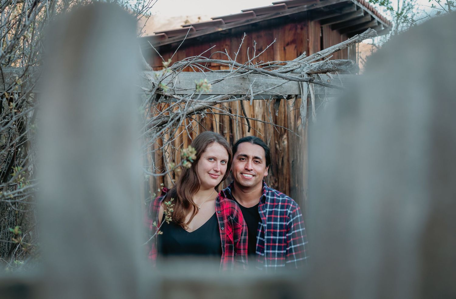 A smiling couple poses together framed by weathered wooden fence posts in front of a rustic barn.