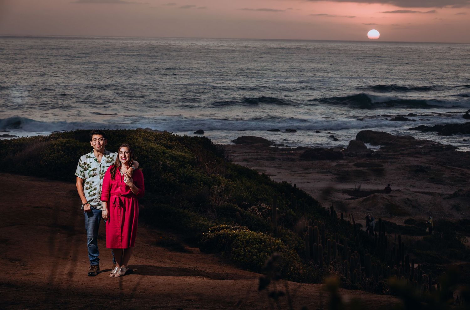 Couple posing at sunset on rocky coastal cliffs with ocean waves and glowing moon in background.