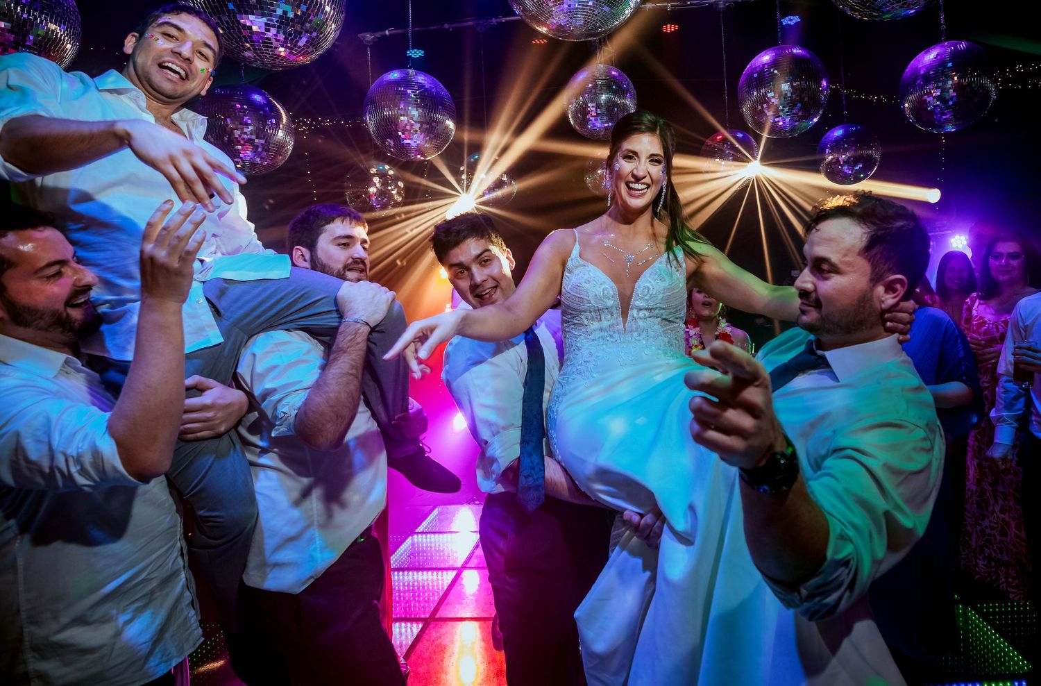 Bride in white gown lifted by groomsmen on a vibrant dance floor with disco balls at a lively wedding reception.