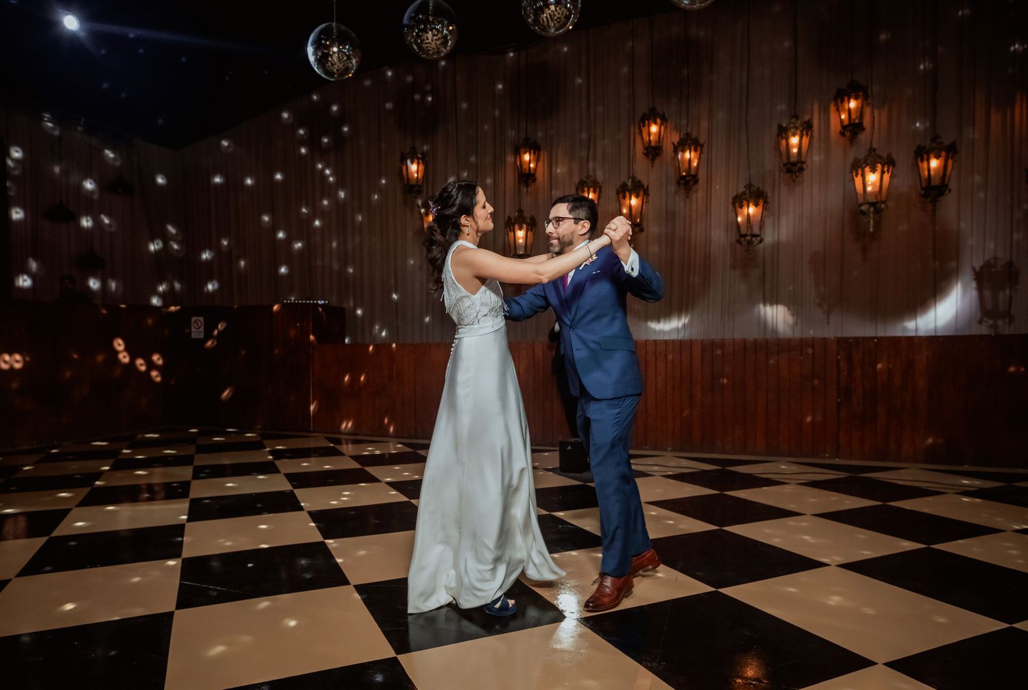 Newlyweds share elegant first dance under disco ball on checkered floor with wall sconces.