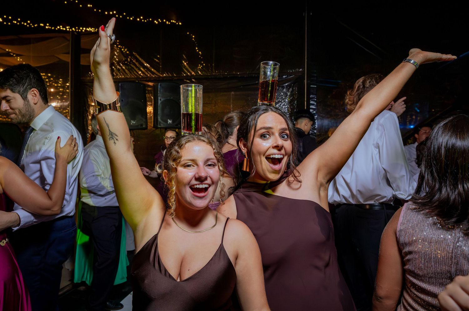 Two joyful women in burgundy dresses celebrating with arms raised at a festive wedding reception.