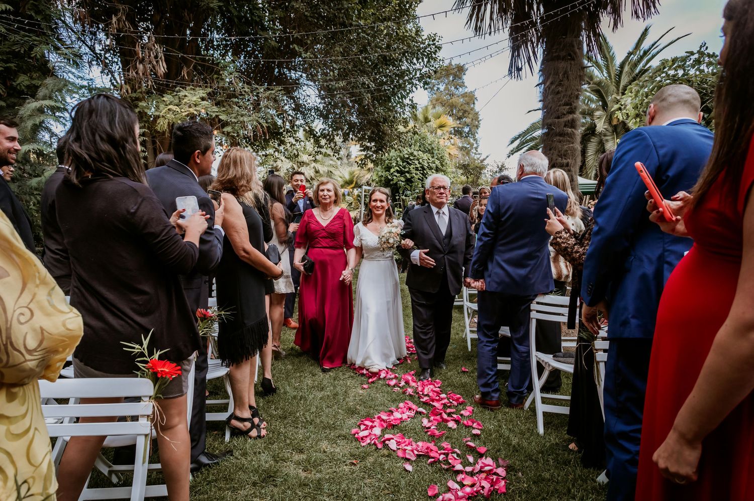 Bride walks down petal-lined aisle with parents through cheering outdoor wedding guests.