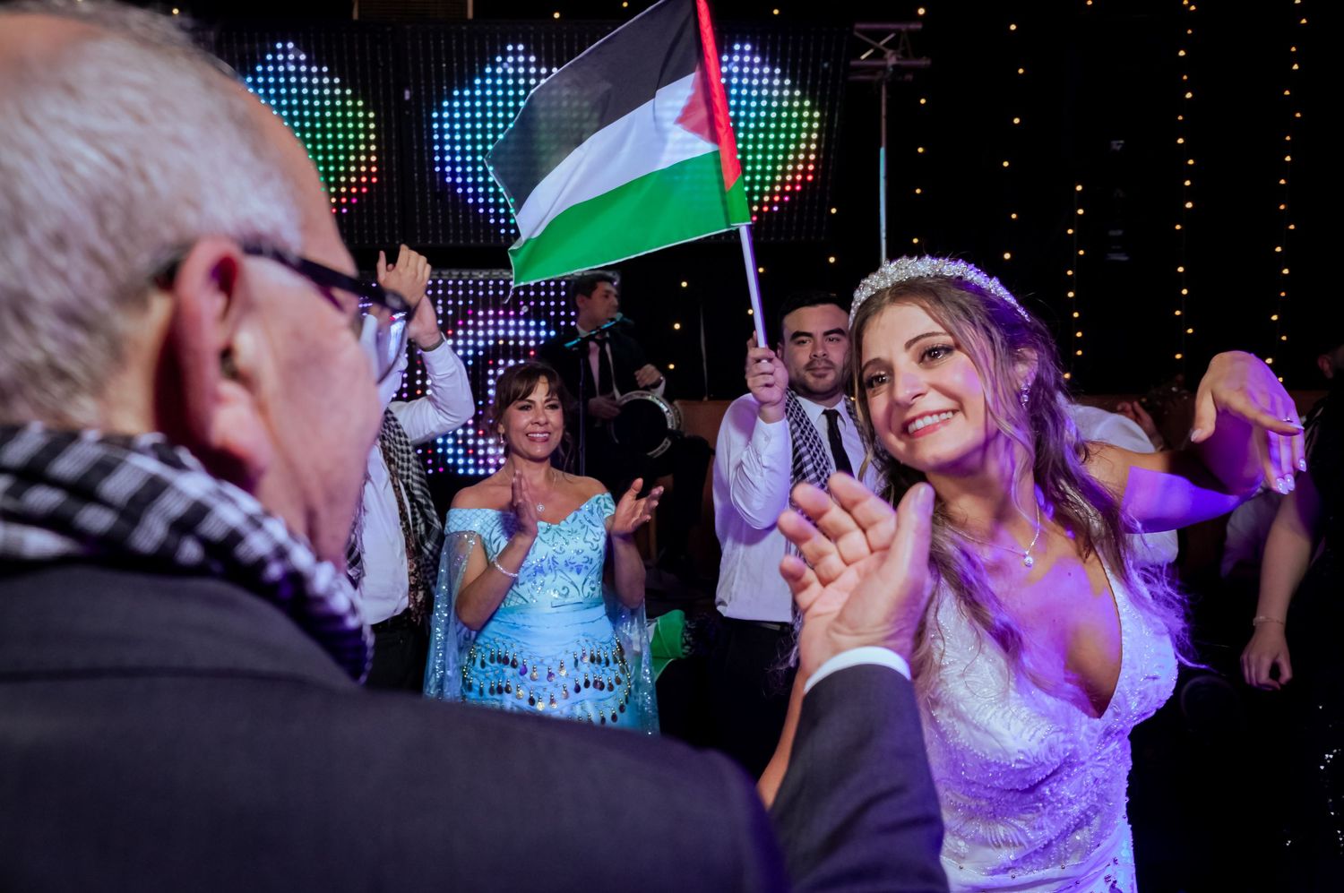 Smiling bride claps with guests waving Palestinian flags during a colorful wedding reception celebration.