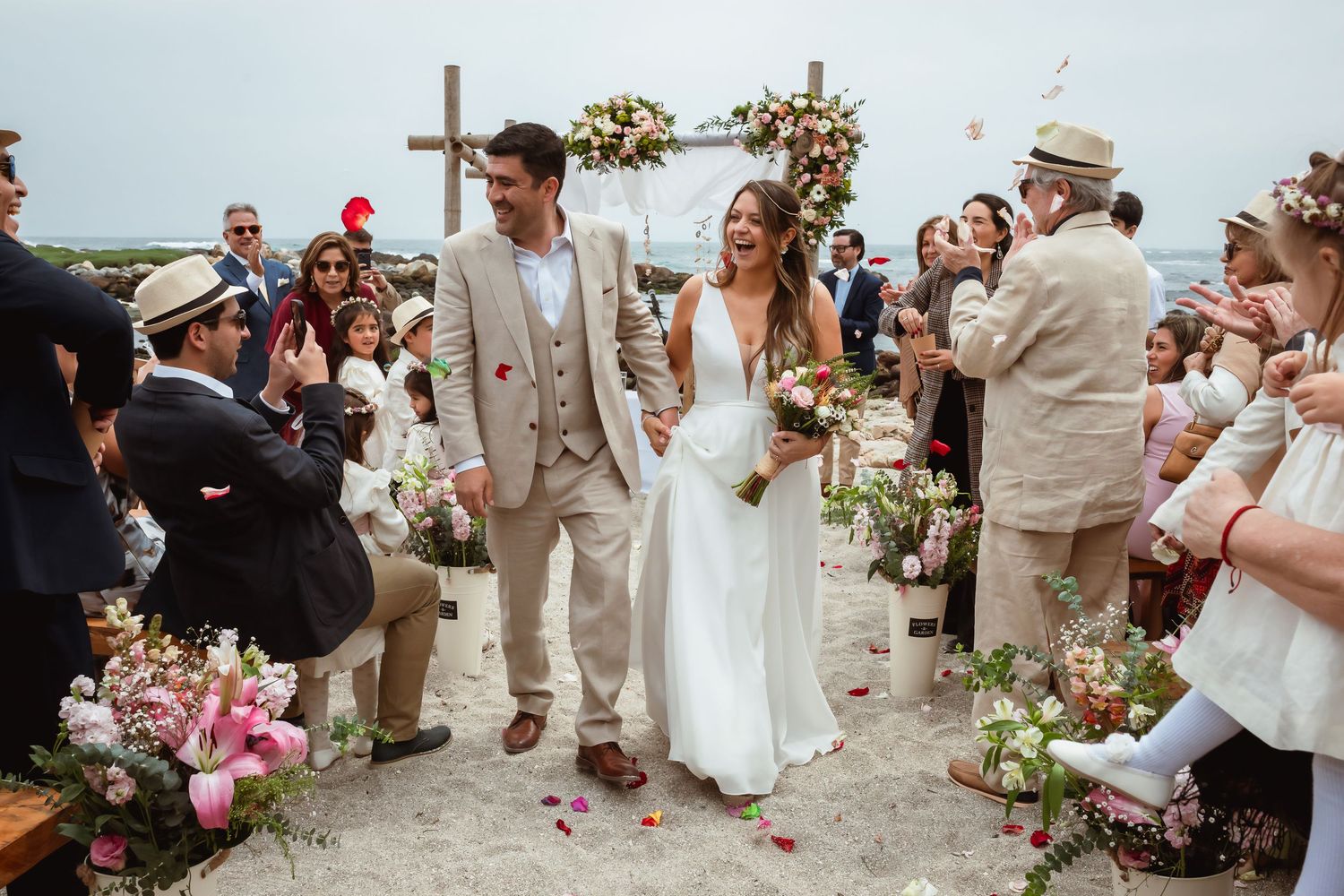 Happy newlyweds walk down the aisle at a beach wedding ceremony surrounded by cheering guests.
