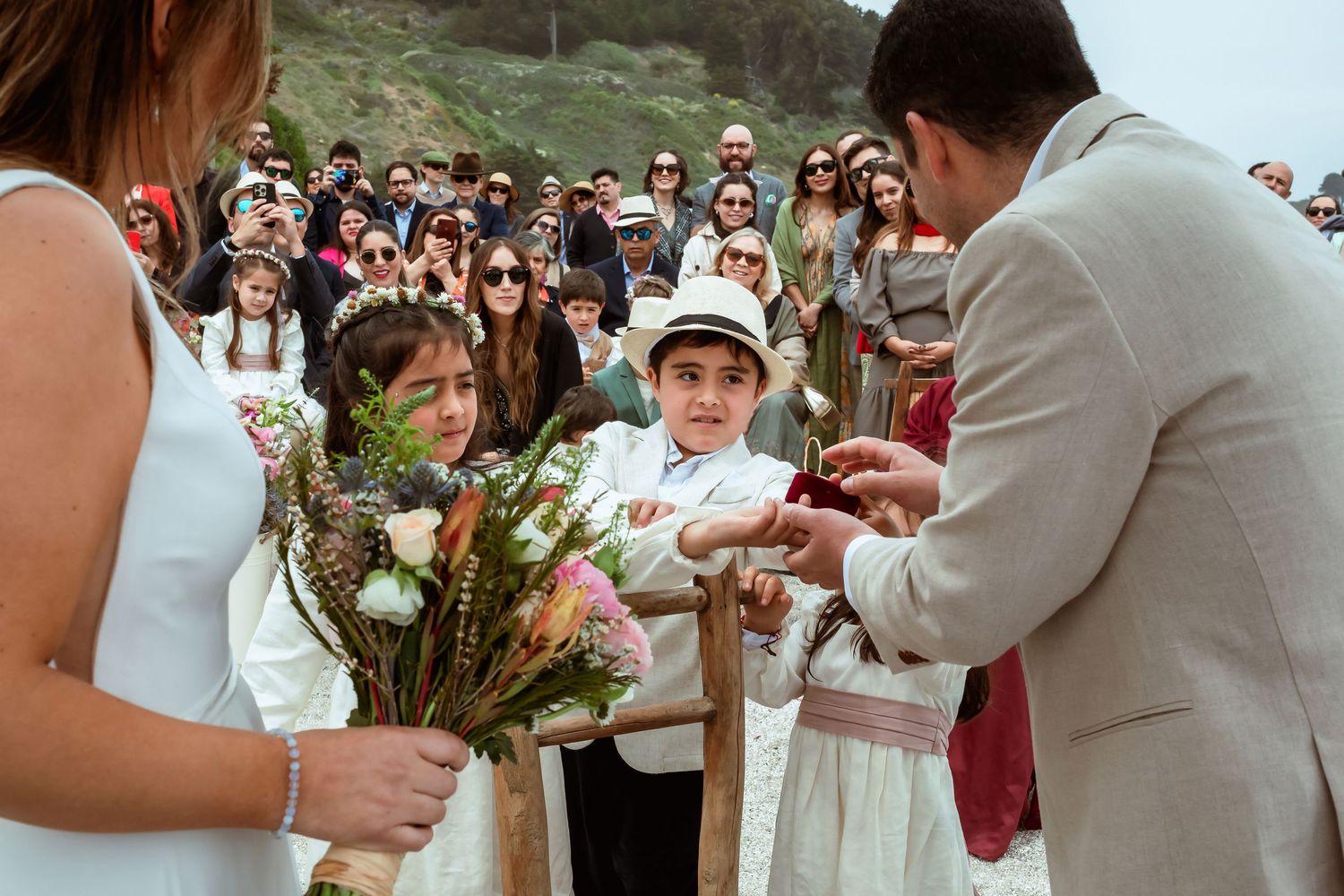 Children present rings to bride and groom during outdoor beach wedding ceremony with guests watching.