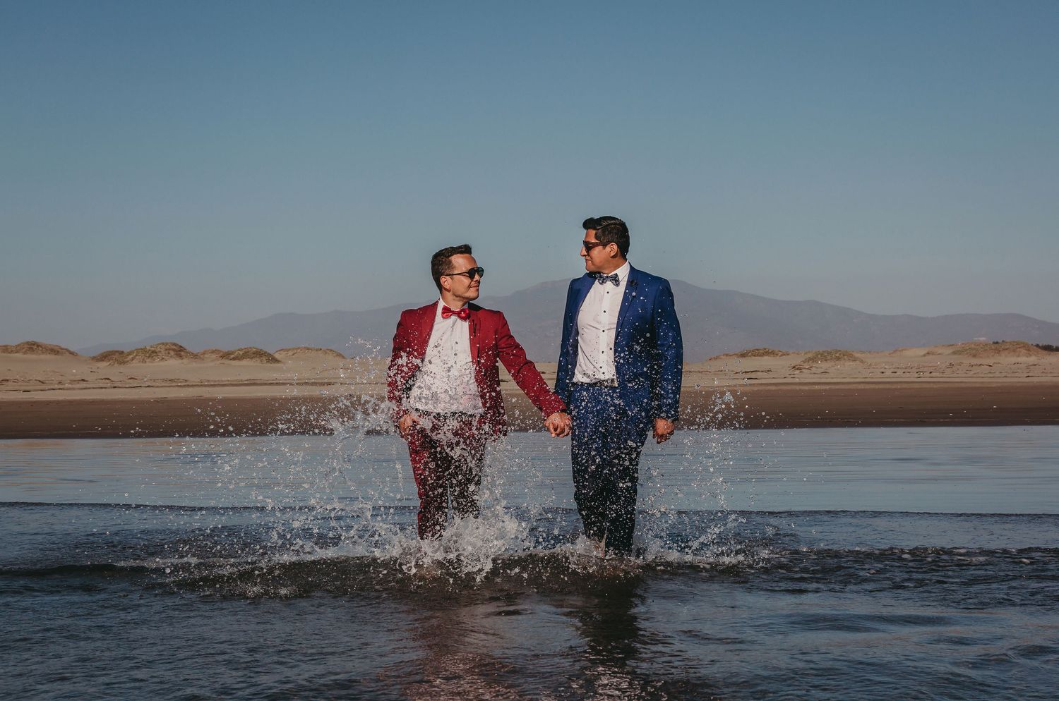 Two grooms in vibrant suits hold hands and gaze at each other while splashing through water.