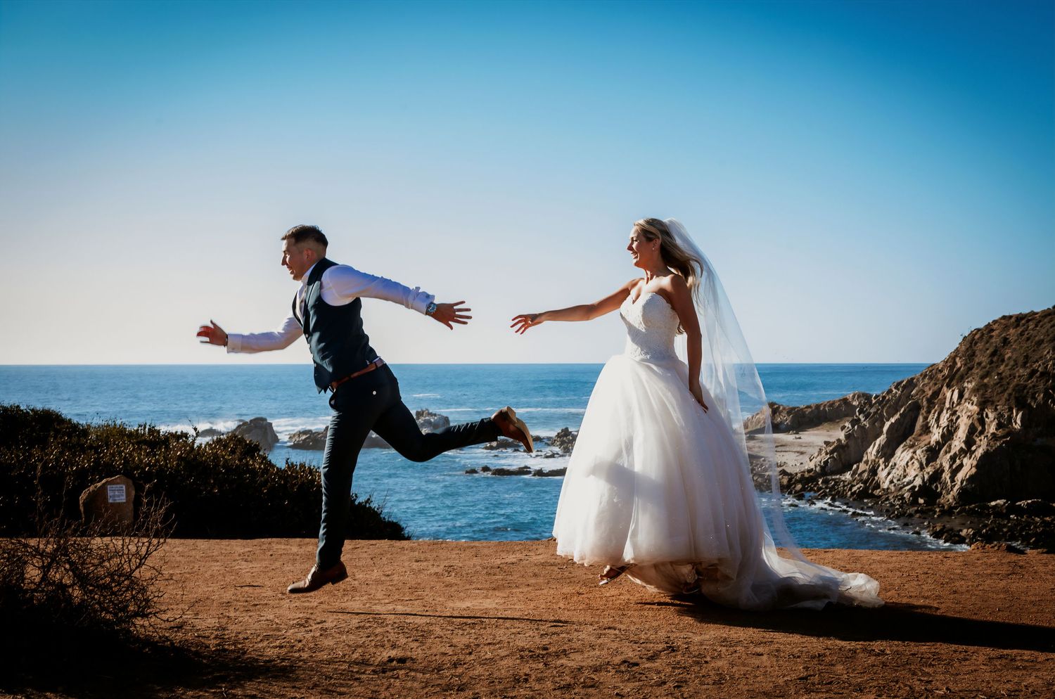Groom joyfully leaps while bride reaches out on a coastal clifftop in wedding attire.