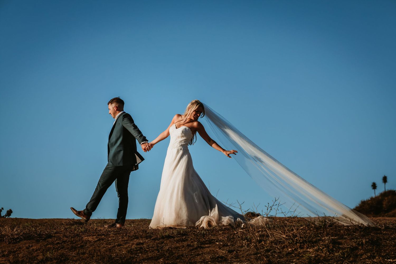 Bride's veil flowing in wind as couple walks hand in hand under clear blue sky.