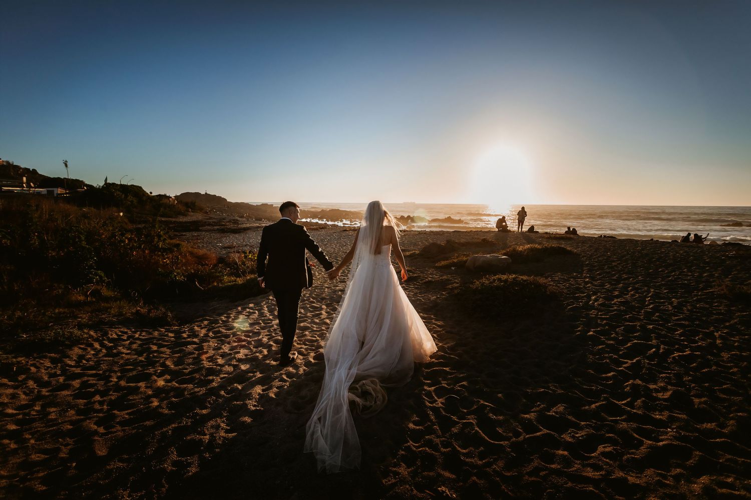 Bride and groom walking hand in hand on sandy beach toward golden ocean sunset.