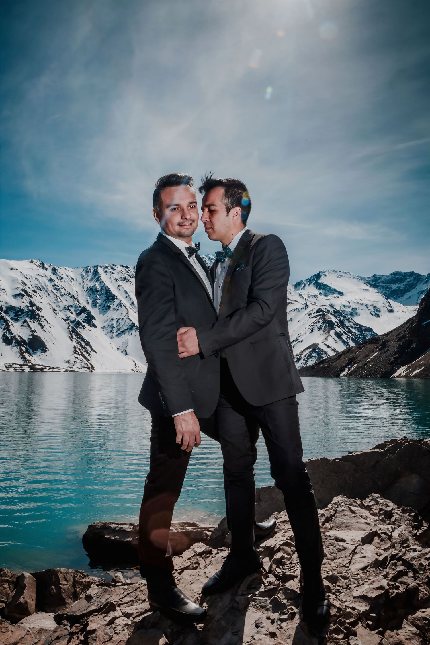 Two grooms share a tender kiss by a glacial lake with dramatic snow-covered mountain scenery.