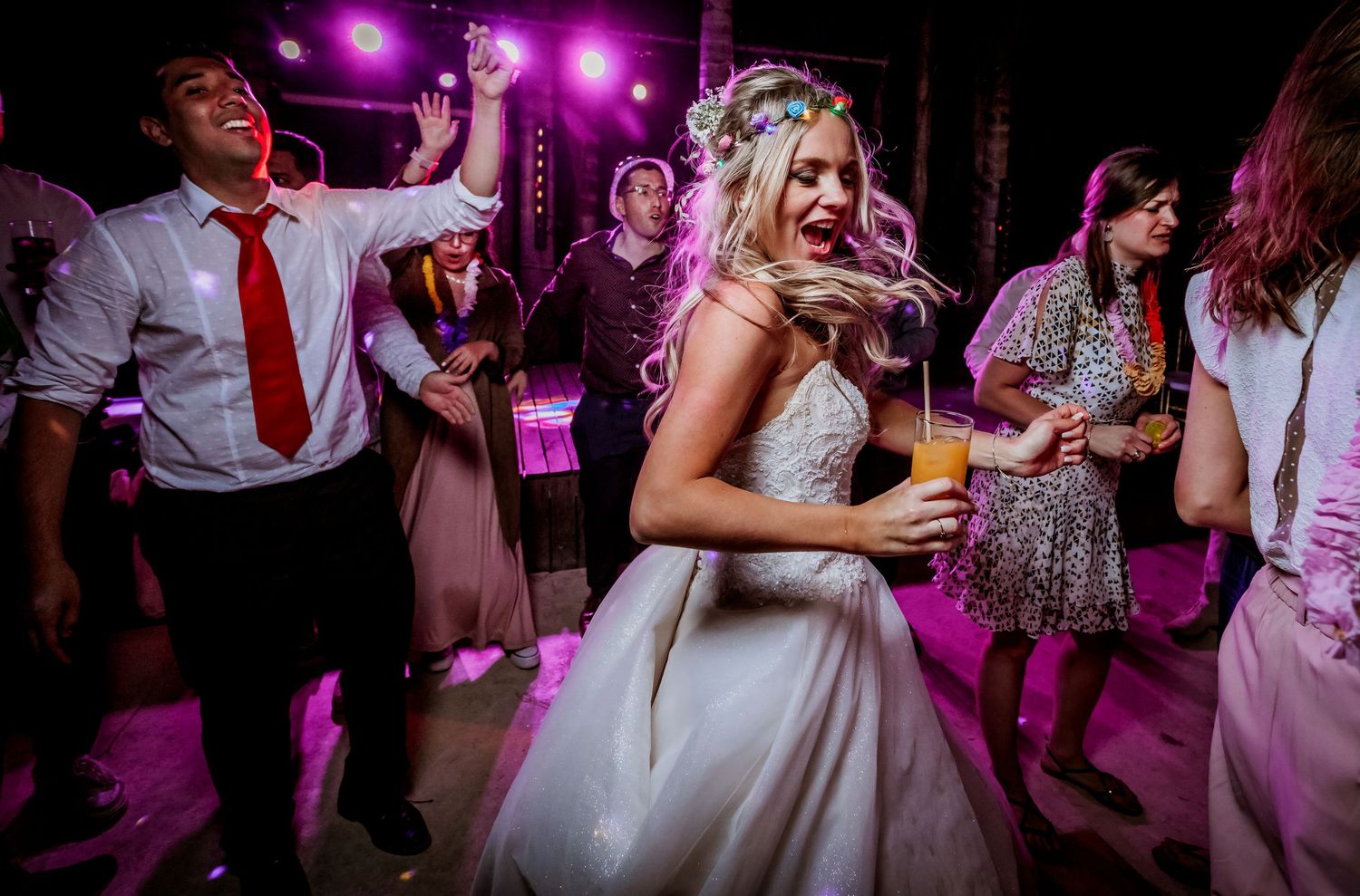 Bride in white gown singing and dancing with guests under colorful wedding reception lights.