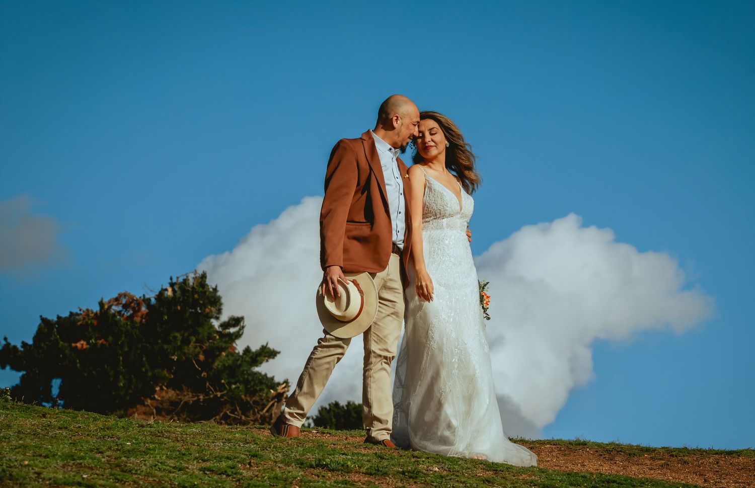 Bride in a white gown and groom in a brown blazer holding a straw hat embrace on a grassy hilltop under a blue sky with white clouds.