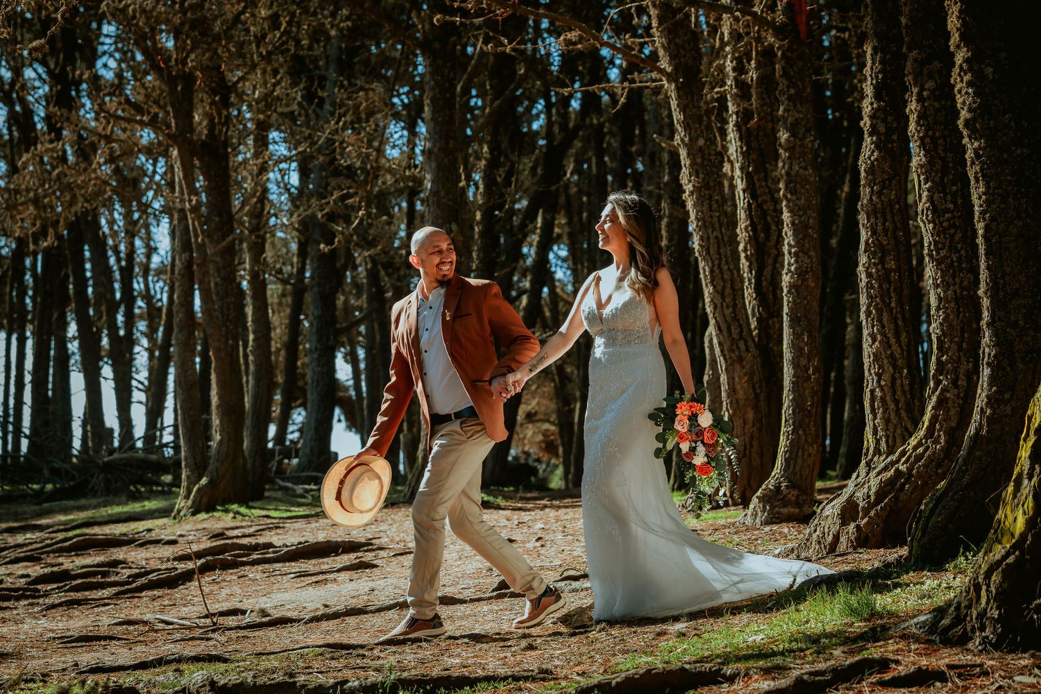 Smiling bride in a white gown and groom in a rust jacket holding a straw hat walk hand in hand through a sunlit pine forest.
