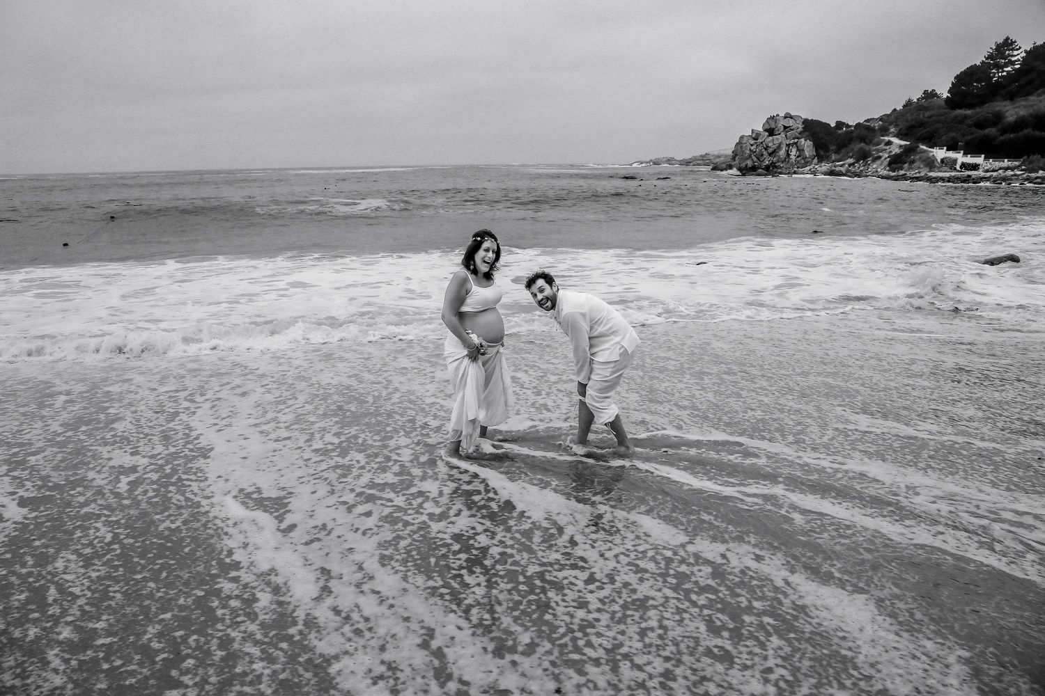 Black and white photo of a couple laughing and playing in ocean waves on a sandy beach.