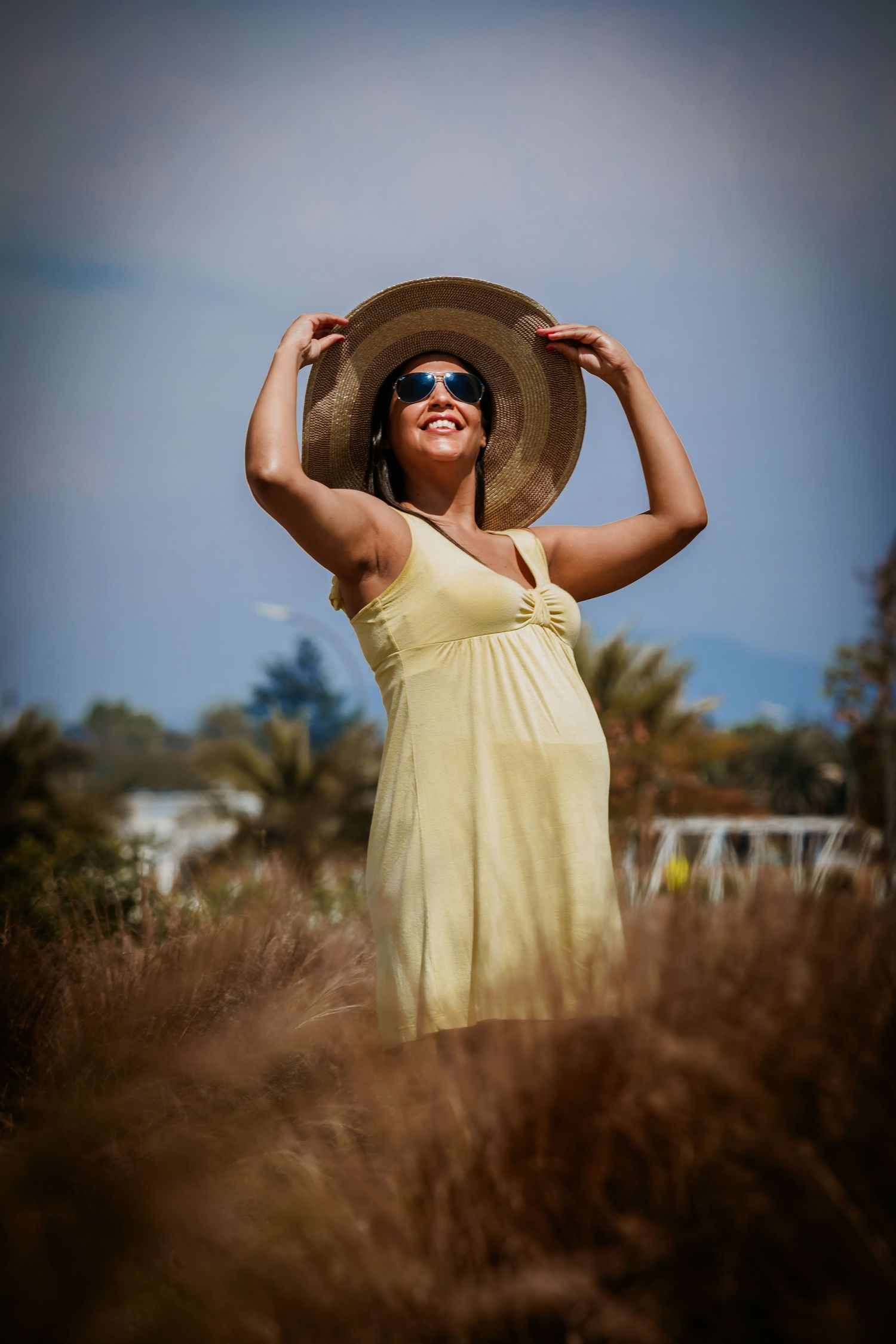 Smiling woman in yellow sundress and wide-brim hat holds her hat outdoors among tall grass with palm trees behind her.
