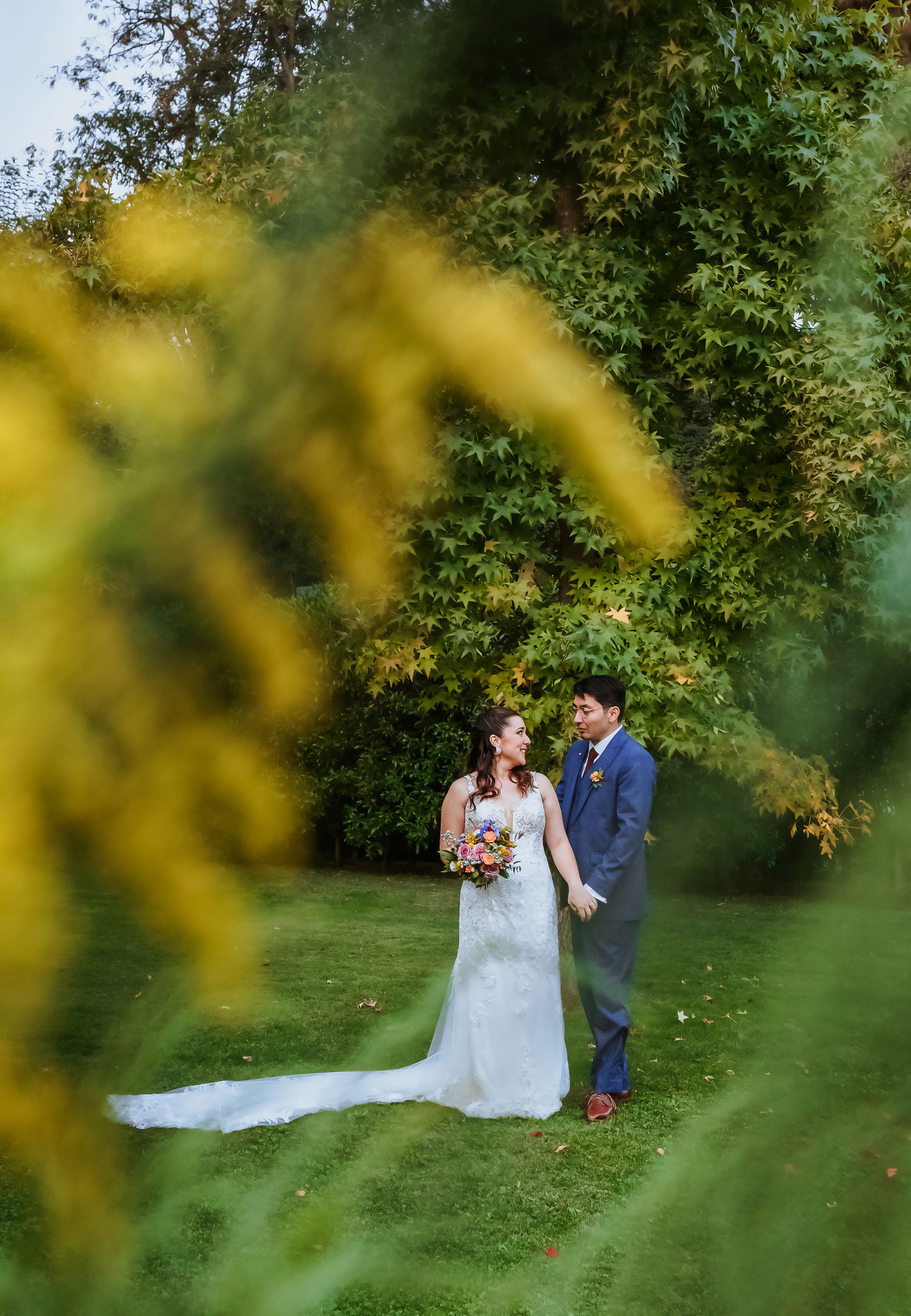 Bride and groom stand together on lush green lawn surrounded by autumn trees with yellow foliage framing the romantic scene.