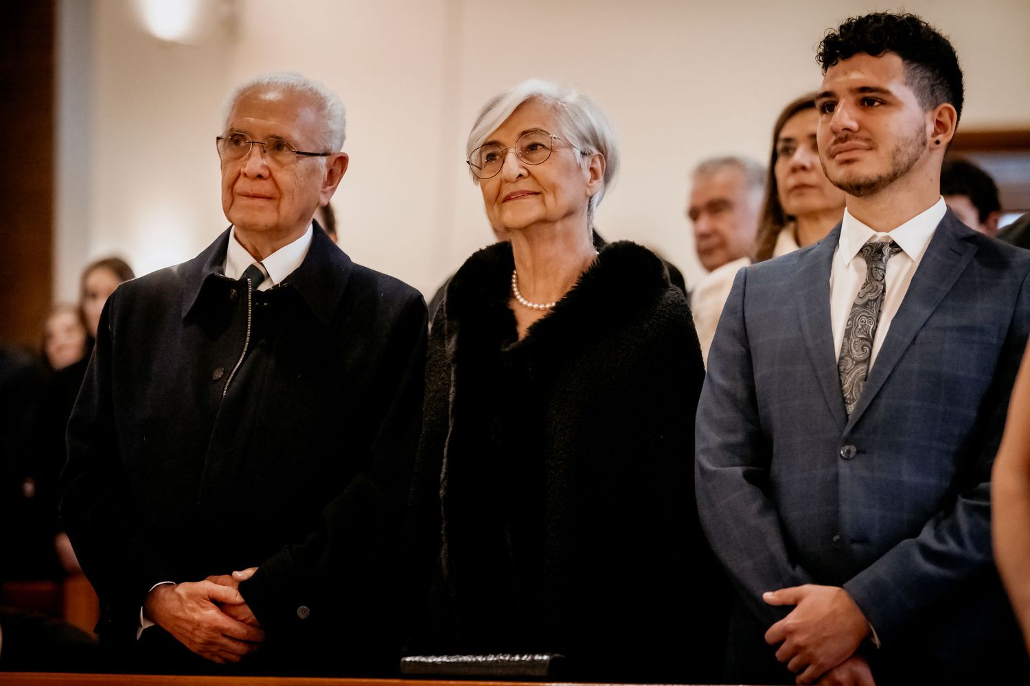 Three people standing solemnly in a church during a formal ceremony, dressed in elegant attire.