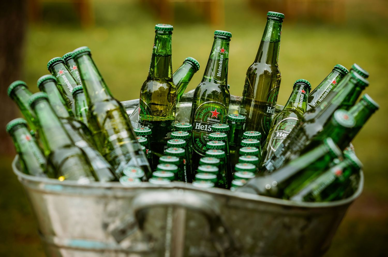 Green beer bottles chilling in an ice-filled metal bucket outdoors at a party or event.